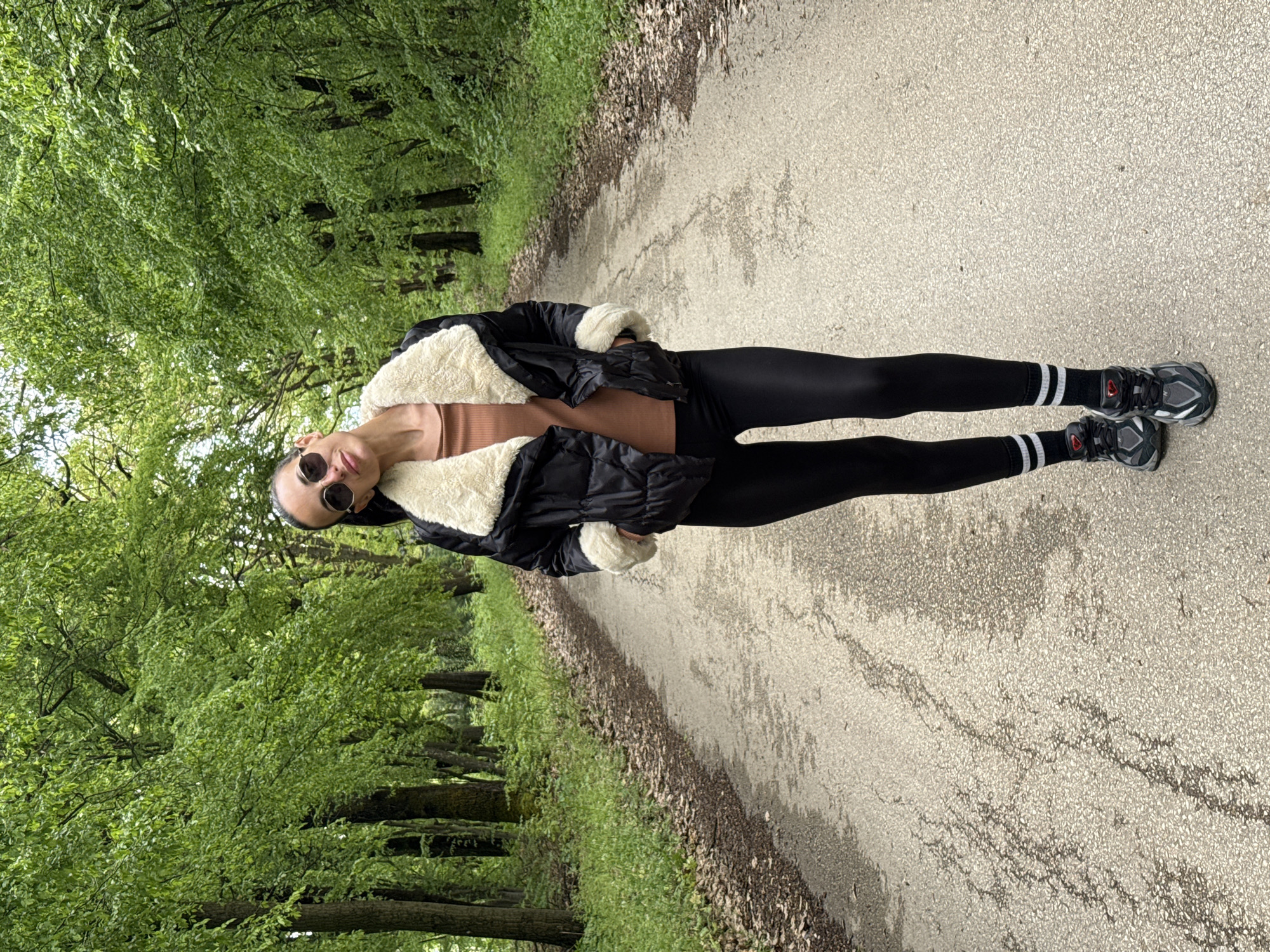 A full-body portrait features a woman standing on a paved path, framed by lush green trees and foliage on both sides. The image is captured from a slightly low angle, looking upwards towards the woman and the dense tree canopy above.
The woman is positioned towards the left of the frame, facing the camera directly. She wears dark sunglasses and has a neutral expression. Her attire includes a black puffer jacket with substantial creamy-white faux fur trim on the collar and cuffs. Beneath the jacket, a fitted nude or light brown top is visible. She is also wearing black athletic leggings, white crew socks with two black stripes near the top, and dark grey athletic sneakers with lighter accents.
The setting appears to be an outdoor park or a tree-lined avenue, identified as being in Pleven, Bulgaria. The paved path extends into the background, flanked by abundant, vibrant green leaves that suggest spring or early summer. The edges of the path show scattered fallen leaves and some natural undergrowth. The lighting is bright and diffused, indicating daytime, possibly under an overcast sky or shaded by the overhead tree canopy, contributing to a soft, natural ambiance. There is no visible text within the image.