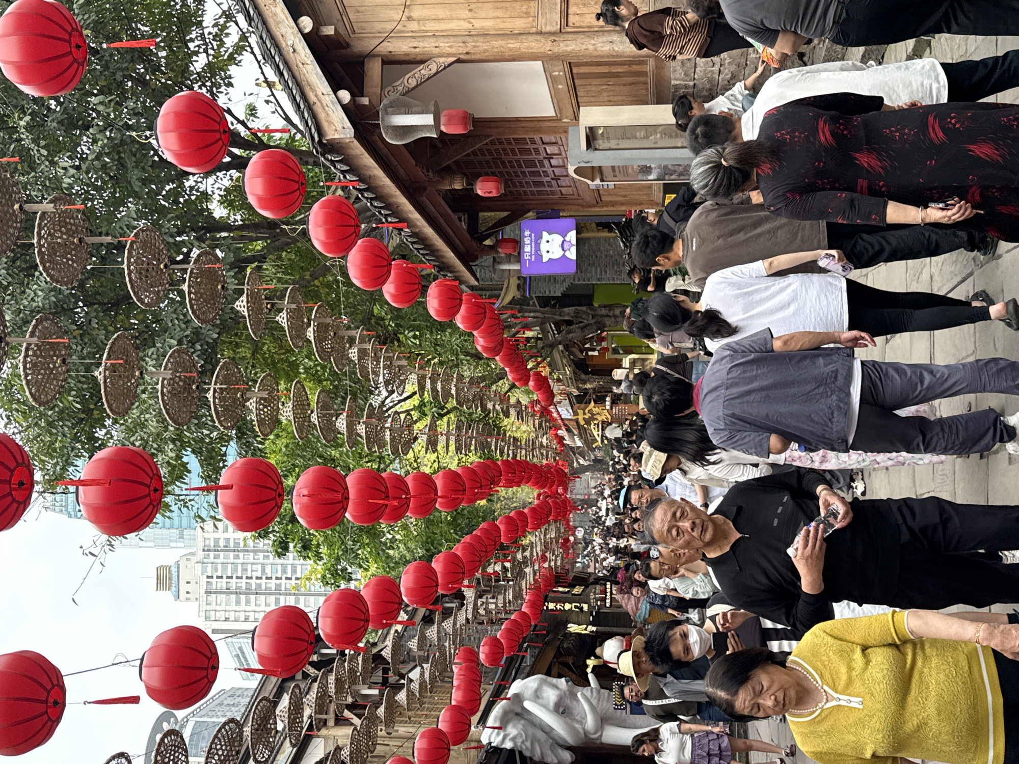 The image captures a bustling pedestrian street scene in Yuzhong District, China. Overhead, rows of red lanterns and woven baskets are suspended along the street's length. The architecture features traditional Chinese-style buildings with wooden facades and overhanging eaves. A large crowd of people, mainly of Asian descent, is visible. Many are dressed in casual attire, and some are looking at their mobile phones. A few people are wearing face masks. A large white sculpture resembling an elephant is located along the street. The environment includes trees, building facades, and a glimpse of modern high-rise buildings in the background.