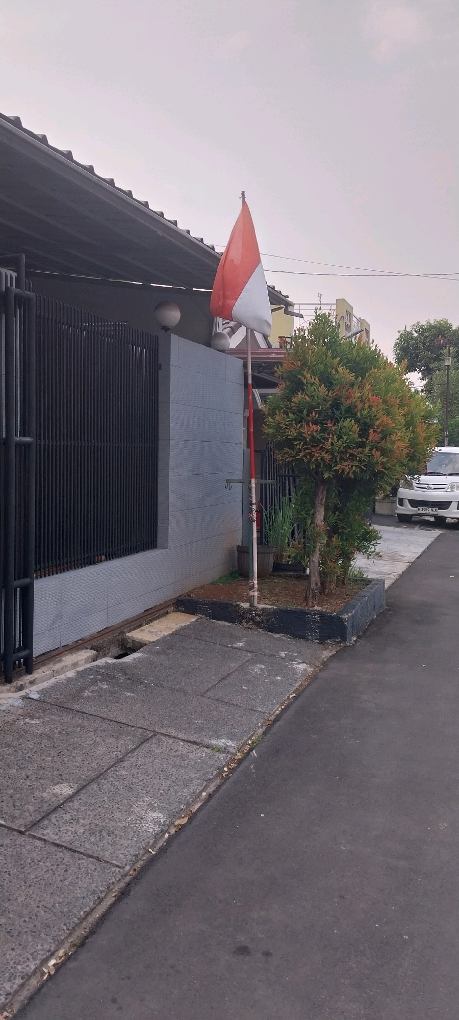 In South Tangerang, Indonesia, an Indonesian flag flies from a pole in front of a grey tiled wall and a black metal fence. The flag, red on top and white on the bottom, is partially obscured by a bushy green tree. Adjacent to the tree is a white van parked on an asphalt road. A paved walkway with a drainage ditch runs alongside the property. The sky is overcast, suggesting a cloudy day.