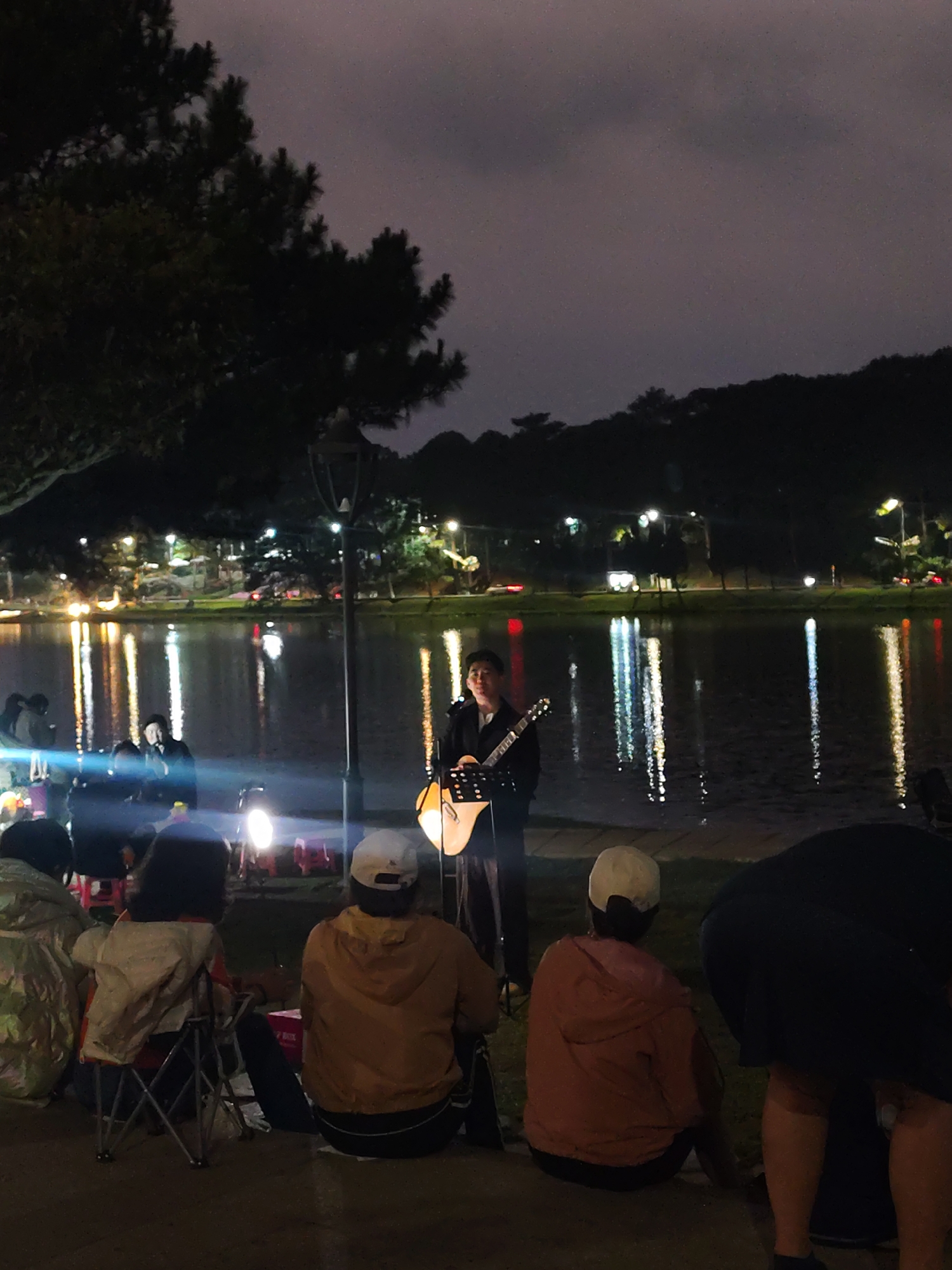 At night, in Da Lat, Vietnam, a man is performing on an electric guitar for a small audience. He is standing by the edge of a body of water, likely Xuan Huong Lake, with trees and streetlights visible across the water. The audience sits on the ground and in folding chairs. There is a lamppost near the performer.