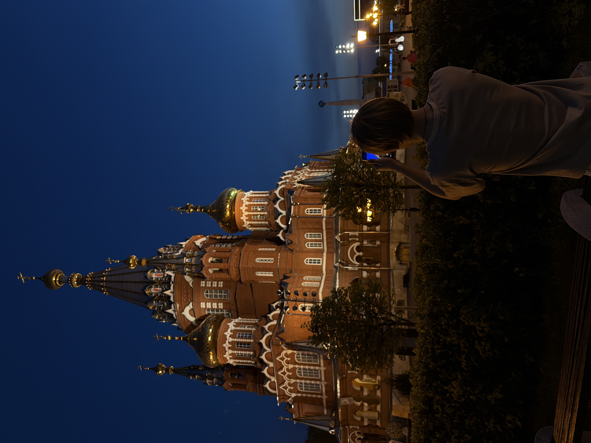 A vertical shot shows a person taking a photograph of St. Michael's Cathedral in Izhevsk, Russia at night. The cathedral, constructed of reddish-brown brick, stands out against a blue sky, with golden domes and intricate white trim highlighting its architectural details. Streetlights, visible above the shrubbery, suggest an urban setting, while the person, shown from the back, is holding a mobile phone and framing the shot of the church. Dark foliage partly obscures the view, and a bench or stone structure is visible in the foreground.