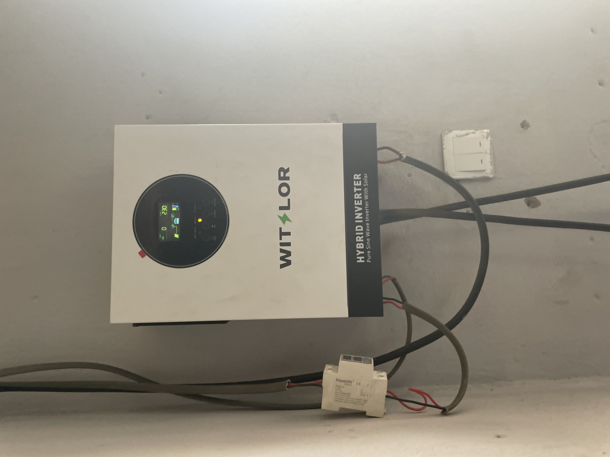 The image shows a wall-mounted hybrid inverter with a circular display screen. The inverter is white with a black frame around the screen and a dark grey section at the bottom. The brand name "WITLOR" is displayed in black letters with a green lightning bolt logo. Text below the brand name reads "HYBRID INVERTER Pure Sine Wave Inverter With Solar". The display screen shows a "0" on the left and "230" on the right, with some icons in between. Various cables, some black and some with red and black wires, are connected to the inverter and run along the wall. A small white electrical component labeled "Posmith" is also attached to the wall near the cables. A double light switch is mounted on the wall to the right of the inverter. The wall appears to be painted a light grey and shows some minor imperfections. The overall scene suggests an electrical installation, likely for solar power, in a residential or commercial setting. The location is identified as Jalingo, Nigeria.