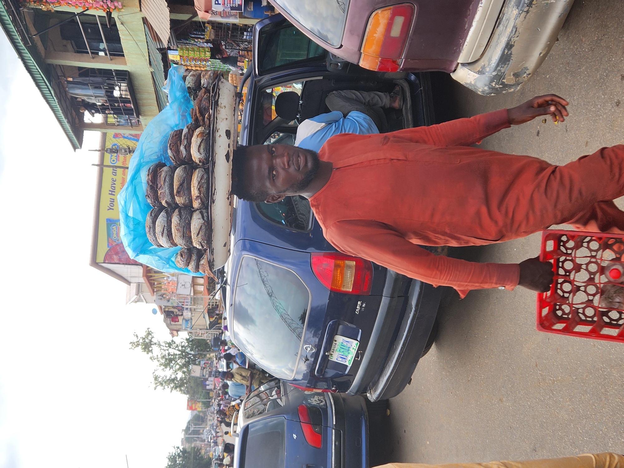 A man stands on a busy street in an urban area of Unknown, Unknown. He is carrying a red plastic crate in his right hand and has a large stack of round, wrapped goods balanced on his head. The goods are covered with blue plastic wrap. The man is wearing a long-sleeved orange shirt. He is positioned near parked cars, including a blue Opel Zafira with a license plate reading GWA-544BK, ABJIA. The street is crowded with people and vehicles. Buildings with signs and shops line the street in the background.
