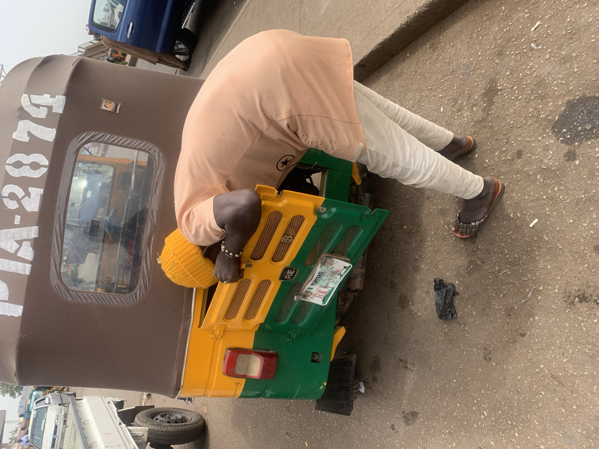 A man in Jos, Nigeria, wearing a yellow knit hat, light peach long-sleeved shirt, and light-colored pants with brown sandals, is bent over the front of a three-wheeled vehicle (tricycle) that is yellow and green. The tricycle’s license plate is damaged and reads "PLATEAU." The back of the tricycle is brown with a number partially visible, reading "DA-287..." Debris sits on the asphalt ground near the tricycle. Other vehicles are visible in the background.
