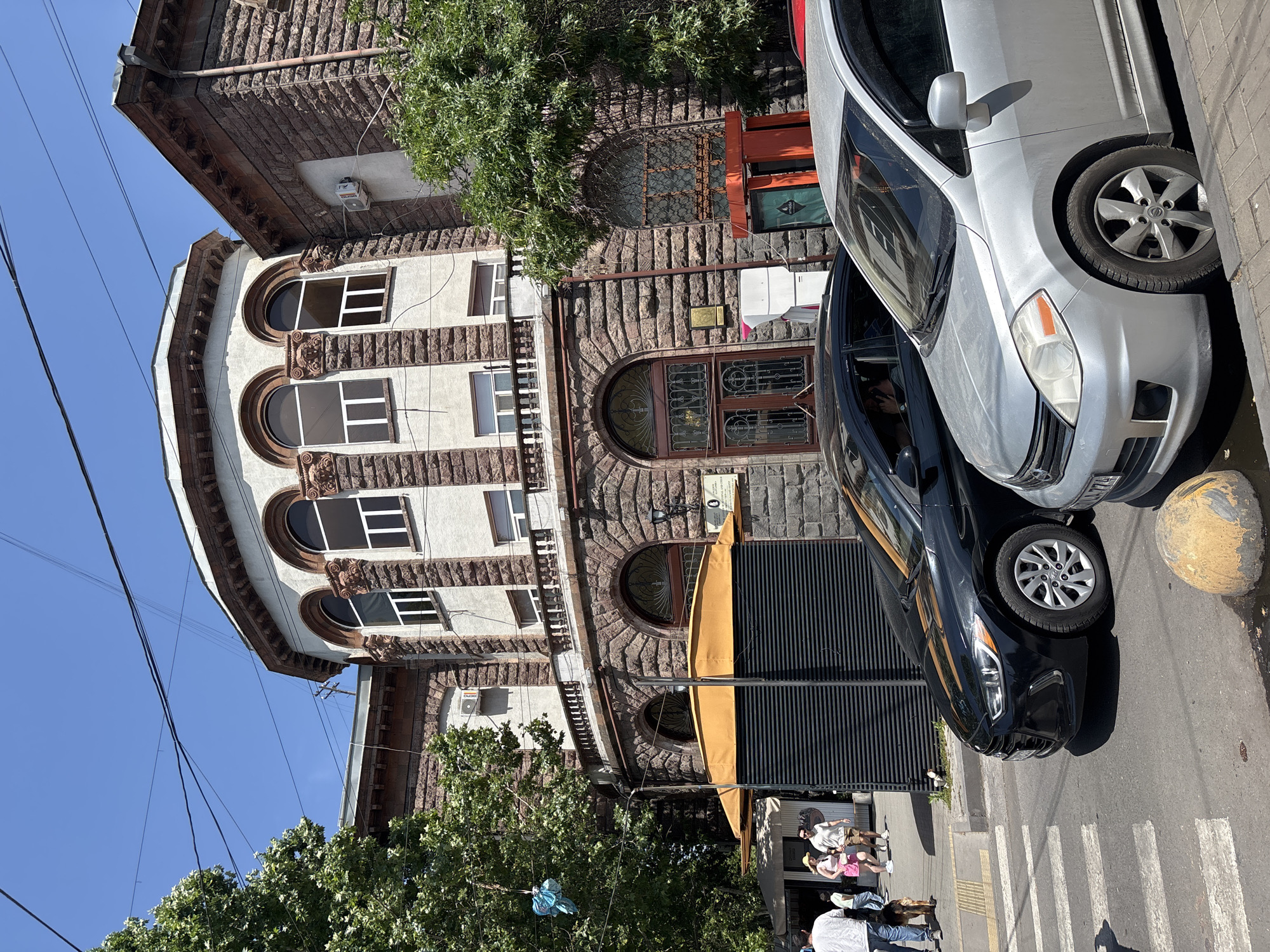 The image shows a street scene in Yerevan, Armenia. In the foreground, a silver car is parked on the right side of the road, with a black car parked to its left. Both cars are facing forward, away from the camera. A pedestrian crossing is visible in the middle of the road.
In the background, there is a large, ornate stone building with arched windows. The building has a rounded section in the center, with a balcony running across the second floor. A yellow awning covers a dark, slatted kiosk in front of the building.
Several people are walking on the sidewalk in front of the kiosk, including a woman in a pink dress and a man in a white shirt and shorts. A dog is also visible on the sidewalk, walking next to a person. Trees with green foliage are planted along the sidewalk.
The sky is clear and blue. The overall impression is of a sunny day in a city with a mix of old and new architecture.