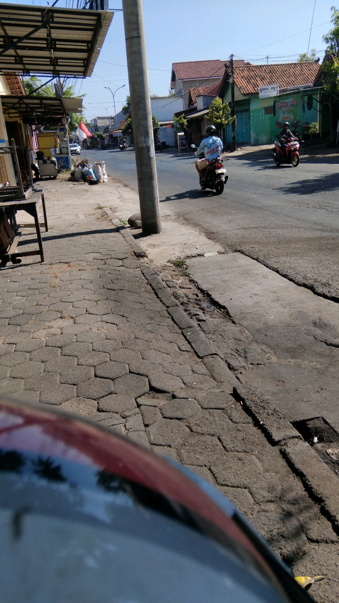 A street scene in Proyonanggan Tengah, Indonesia features a busy road with several people on motorbikes. The foreground is dominated by the blurred reflection of a vehicle, obscuring the view of what appears to be the driver's perspective. To the left, a sidewalk made of hexagonal pavers runs alongside a street-side stall. Further down the street, a concrete utility pole stands tall, casting a shadow on the road. Two men on motorbikes are visible, one in the middle ground and another further in the background. The man in the middle ground, closer to the camera, is wearing a light-colored shirt with a graphic print and is riding a white motorbike. The man in the background is on a red motorbike. Buildings with orange tiled roofs line the street, and lush green trees provide a natural element to the urban landscape. The sky is clear and bright, indicating a sunny day. Several bags of what appear to be merchandise or waste are piled up near the stall. The overall atmosphere is that of a typical Indonesian street scene.