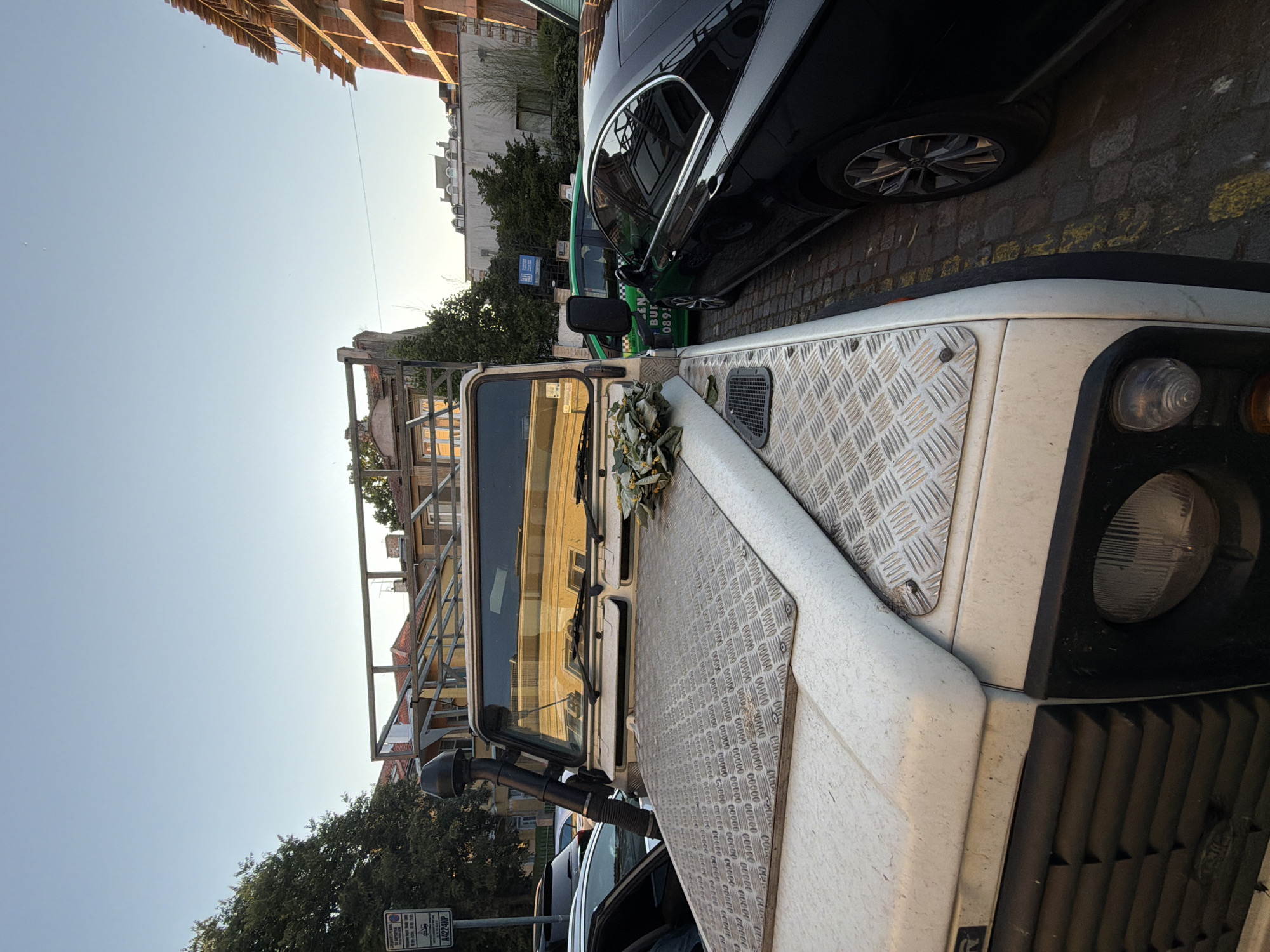 The image shows a white Land Rover Defender parked on a cobblestone street. The vehicle has a snorkel on the driver's side and a metal cage-like structure over the roof. There is a bunch of dried leaves and flowers placed on the hood. A black car is parked to the right of the Land Rover, and a green taxi is visible behind it. In the background, there are buildings under construction and older residential buildings. A street sign with Bulgarian text and a license plate that reads "A422KP" is visible on the left side of the image. The scene is set in the city of Burgas, Bulgaria, during daylight hours, as indicated by the clear sky and shadows.