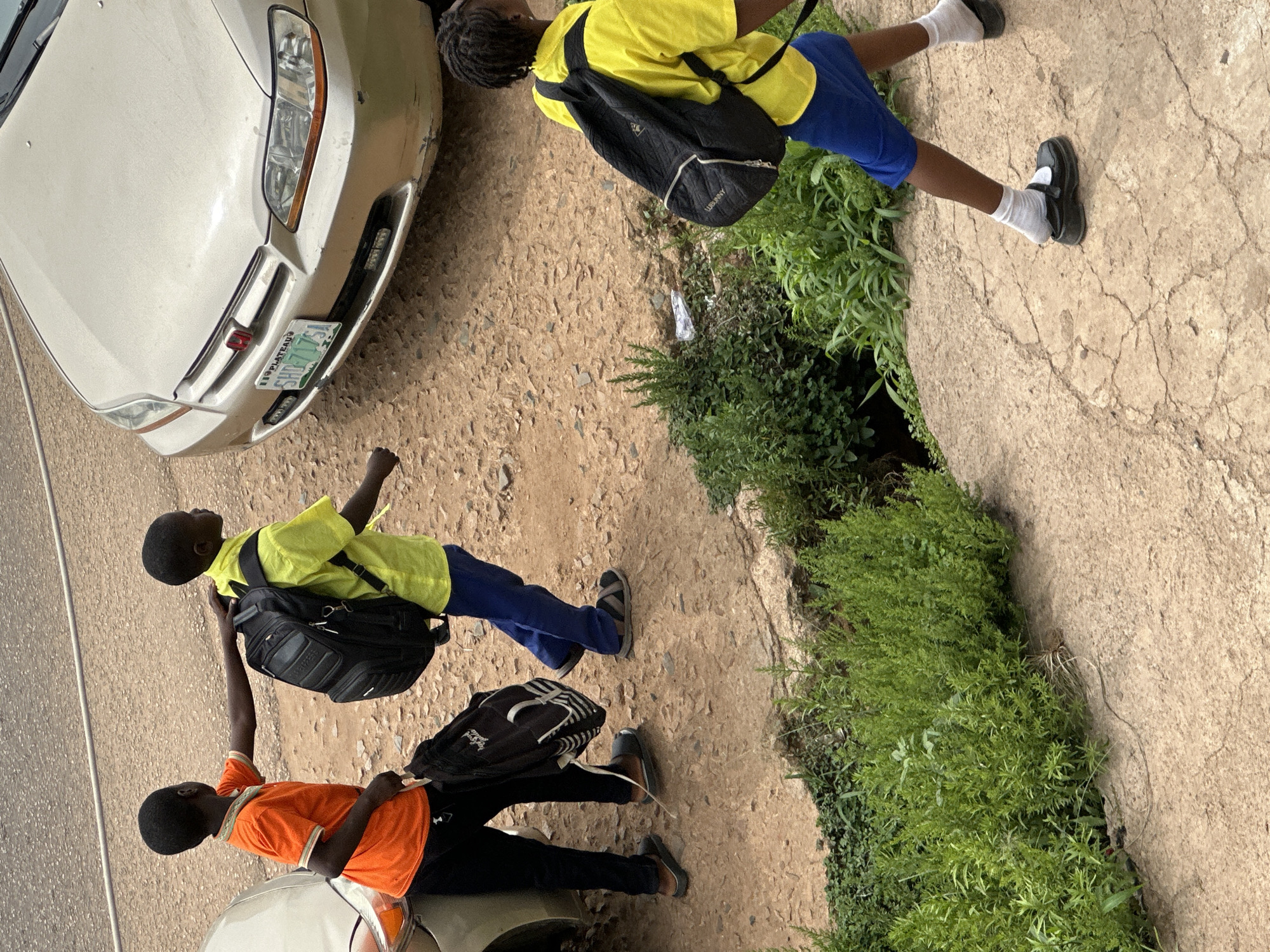 Two young boys, wearing school uniforms of orange and yellow shirts respectively, walk along a dirt path in Jos, Nigeria. The boy in the orange shirt has his hand on the backpack of the boy in the yellow shirt, who carries a large black backpack. They are walking past a parked gold sedan. Further down the path, another child in a yellow shirt and blue shorts, also wearing a backpack, walks away from the camera. The area is characterized by sparse vegetation and a rough, uneven ground surface.