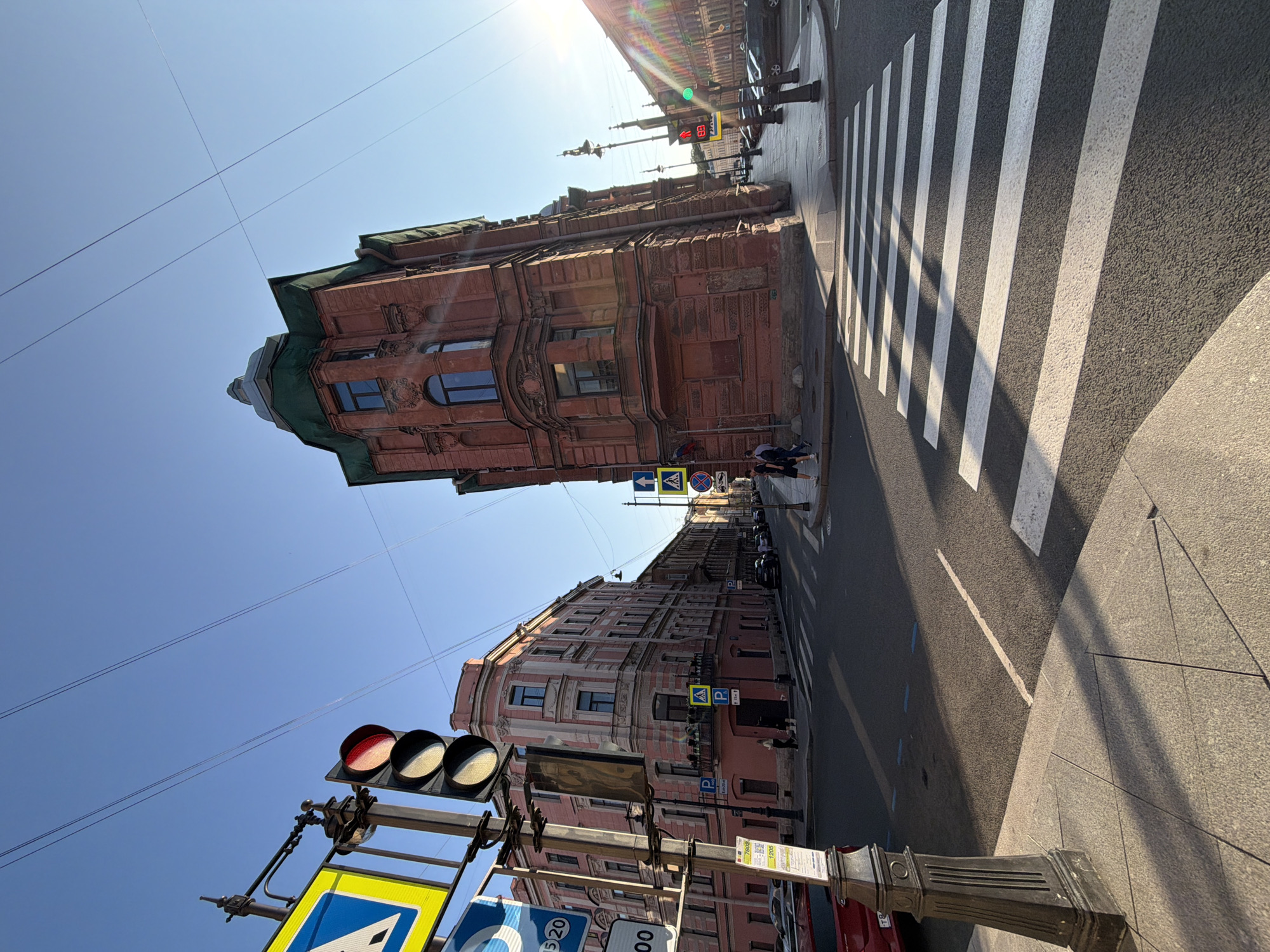 This is an outdoor, street-level, wide-angle photograph taken in Saint Petersburg, Russia, on a sunny day. The viewpoint is from a sidewalk looking across an intersection.
In the foreground, a pedestrian crosswalk with white stripes is visible on the asphalt road. To the left, a black, ornate lamppost stands on the granite sidewalk, casting a long shadow. Adjacent to the lamppost, a traffic light with a red light illuminated is attached to a metal pole, which also holds several road signs, including blue and yellow warning signs with white symbols and a blue sign with white text and numbers.
Dominating the mid-ground and right side of the image is a large, reddish-brown brick building with green architectural elements on its roof and windows. It has a distinct corner structure and detailed stonework around its windows and at the base. To the left of this building, a narrower, lighter pink building with multiple floors and ornate details is visible.
The intersection is clear of vehicles at the immediate crossing point. In the background, more buildings line the street, and parked cars are visible along the curb. Several pedestrians are seen walking on the sidewalks and near the crosswalks. The sky is clear and blue, with power lines crisscrossing overhead. The sun is low in the sky, casting long shadows and creating lens flare on the right side of the image. The overall scene depicts a typical urban street corner in Saint Petersburg, Russia.