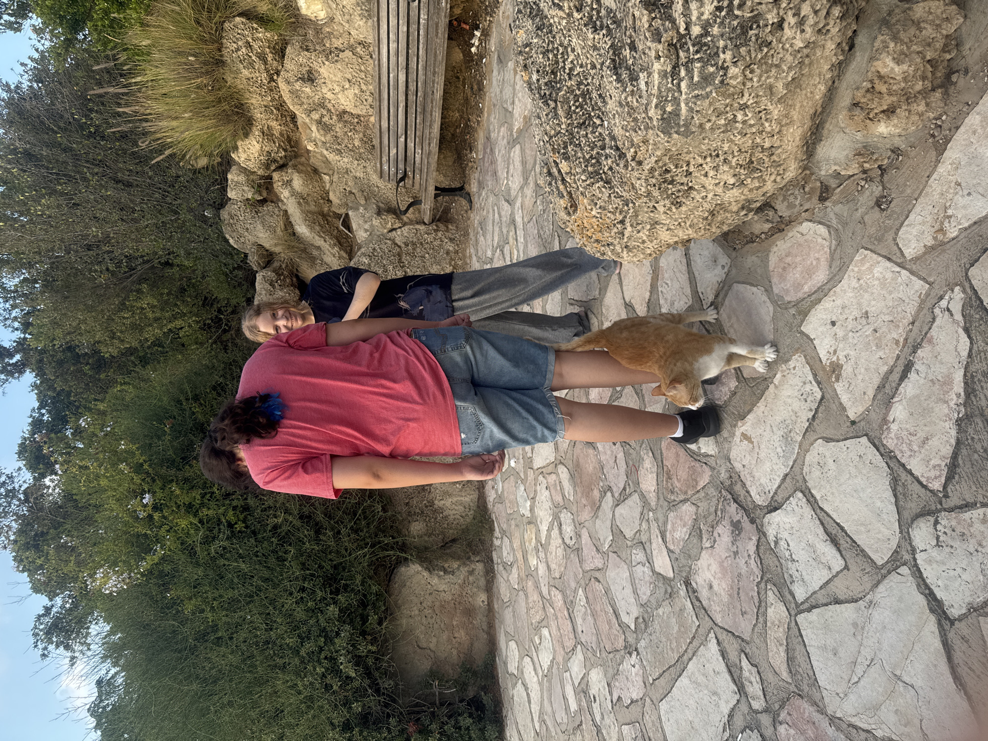 A ginger and white cat stands on a stone pathway in front of two people. The person on the left, facing away from the camera, wears a bright pink t-shirt and denim shorts. Their hair is dark and appears to have blue streaks. The person on the right, facing the camera and smiling, wears a dark t-shirt and grey sweatpants. Behind them, a wooden bench is situated against a rocky embankment with lush green bushes and tall grasses. The scene is set in a park-like outdoor environment, likely within the city of Ramat Gan, Israel, given the context. The lighting suggests it is late afternoon or early evening.