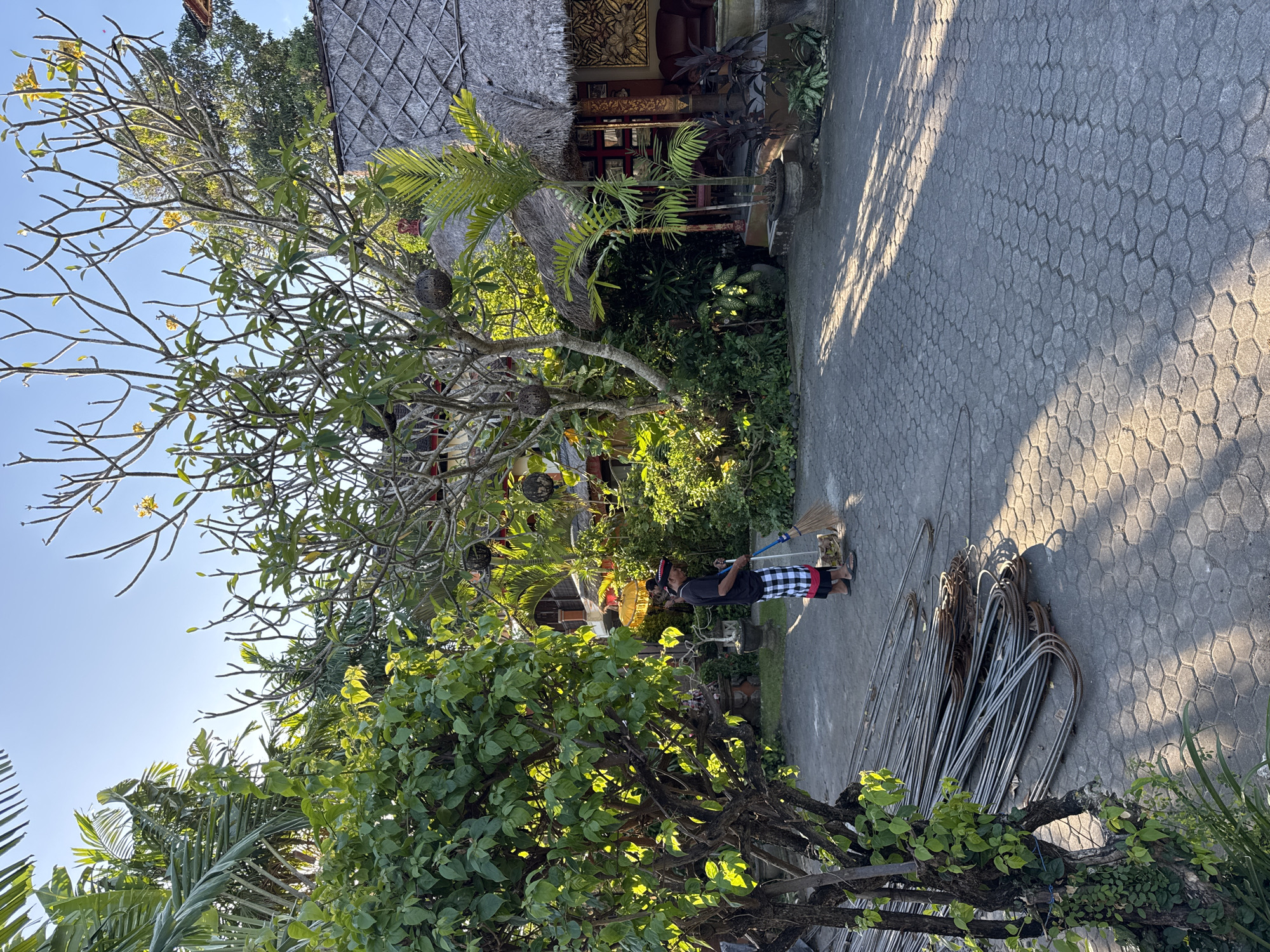 A man sweeps in Kuta, Indonesia. The man is wearing a black t-shirt, checkered shorts, and sandals. He is holding a broom and sweeping debris on a paved surface. Next to the man is a pile of curved metal rods. A building with a thatched roof and intricate carvings is visible in the background, along with lush tropical vegetation. The scene is illuminated by sunlight, casting shadows on the ground.