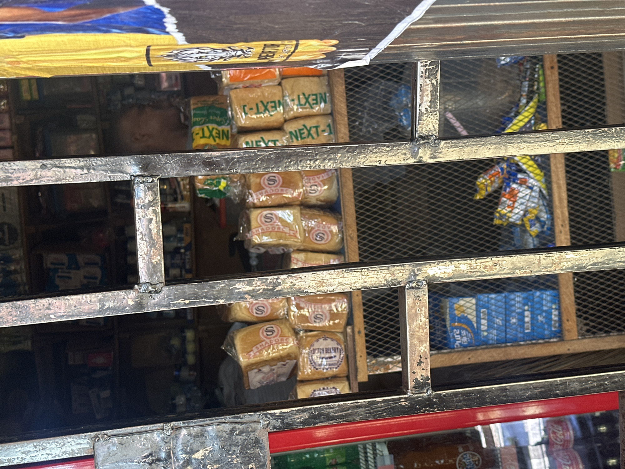 The image depicts a partial view into a small, informally structured shop or kiosk, viewed through a heavily rusted and worn metal security gate. The location is in the city of Unknown, Unknown.
In the immediate foreground, a dark, patinated metal security gate, constructed of welded horizontal and vertical bars, occupies the central frame. The metal shows significant wear, with chipped paint and visible rust. To the far left, a section of a red metal frame is visible, likely part of the shop's entrance structure.
Through the gate, the interior of the shop is visible, appearing somewhat dimly lit. On an upper wooden shelf, multiple commercially packaged loaves of bread are stacked. Some packages bear a prominent 'S' logo and the brand "SHADORE", while others feature green text reading "NEXT-J" or "NEXT". Behind these bread loaves, the indistinct head and shoulder profile of a person, possibly a shopkeeper or customer, is faintly visible, facing towards the right side of the frame.
Below the bread, additional wooden shelving is present, and lower sections of the shop feature a dark metal mesh or chicken wire acting as a barrier or part of the shelving system. On a lower shelf, a stack of blue boxes, possibly "Pure Bliss" brand, is visible. Scattered, colorful plastic bags, likely containing snacks, are also discernible on a shelf near the bottom of the frame. The background of the shop reveals further shelves stocked with various, largely indistinct items.
To the far left of the image, partially obscured by the red frame, a refrigerated display unit is visible, showing bottles with "Coca-Cola" and "Fanta" branding. On the far right, a vertical poster or banner is partially visible, featuring bright yellow and blue sections, with a white and black graphic (possibly an animal or tribal motif) and partial text reading "PERITO". Adjacent to the poster on the right is a vertical section of corrugated metal, suggesting an exterior wall or another adjacent structure.