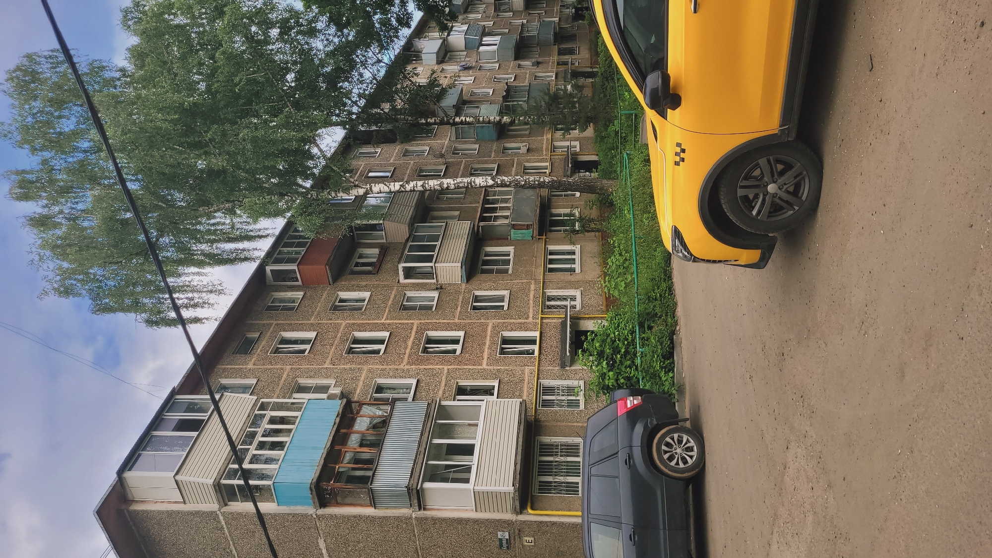 The image shows a street scene in front of a multi-story apartment building with balconies. The building has a concrete facade and appears to be in a residential area. There are trees and greenery surrounding the building. In the foreground, a yellow taxi and a dark SUV are parked on a dirt road. The sky is overcast with clouds. A street sign with Cyrillic letters is visible on the building. The overall impression is a typical urban landscape.