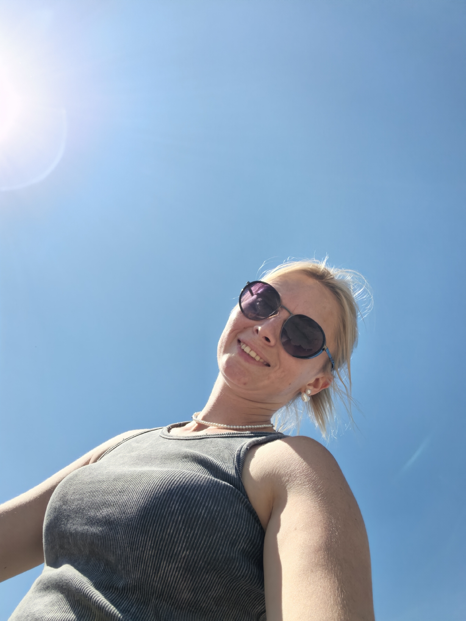 A low-angle, outdoor, medium shot captures a blonde woman against a clear blue sky with the sun glinting on the left side of the frame. She is wearing a grey ribbed tank top, a pearl necklace, and large, round, dark-lensed sunglasses. The woman's head is tilted back and she is smiling. Her fair skin is visible, with freckles across her nose and cheeks. Her blonde hair is pulled back, with a few strands loose around her face. A pearl earring is visible on her right ear. The overall setting appears to be sunny and open. The image does not contain any elements that would allow for identification of a specific location within Sterlitamak, Russia.