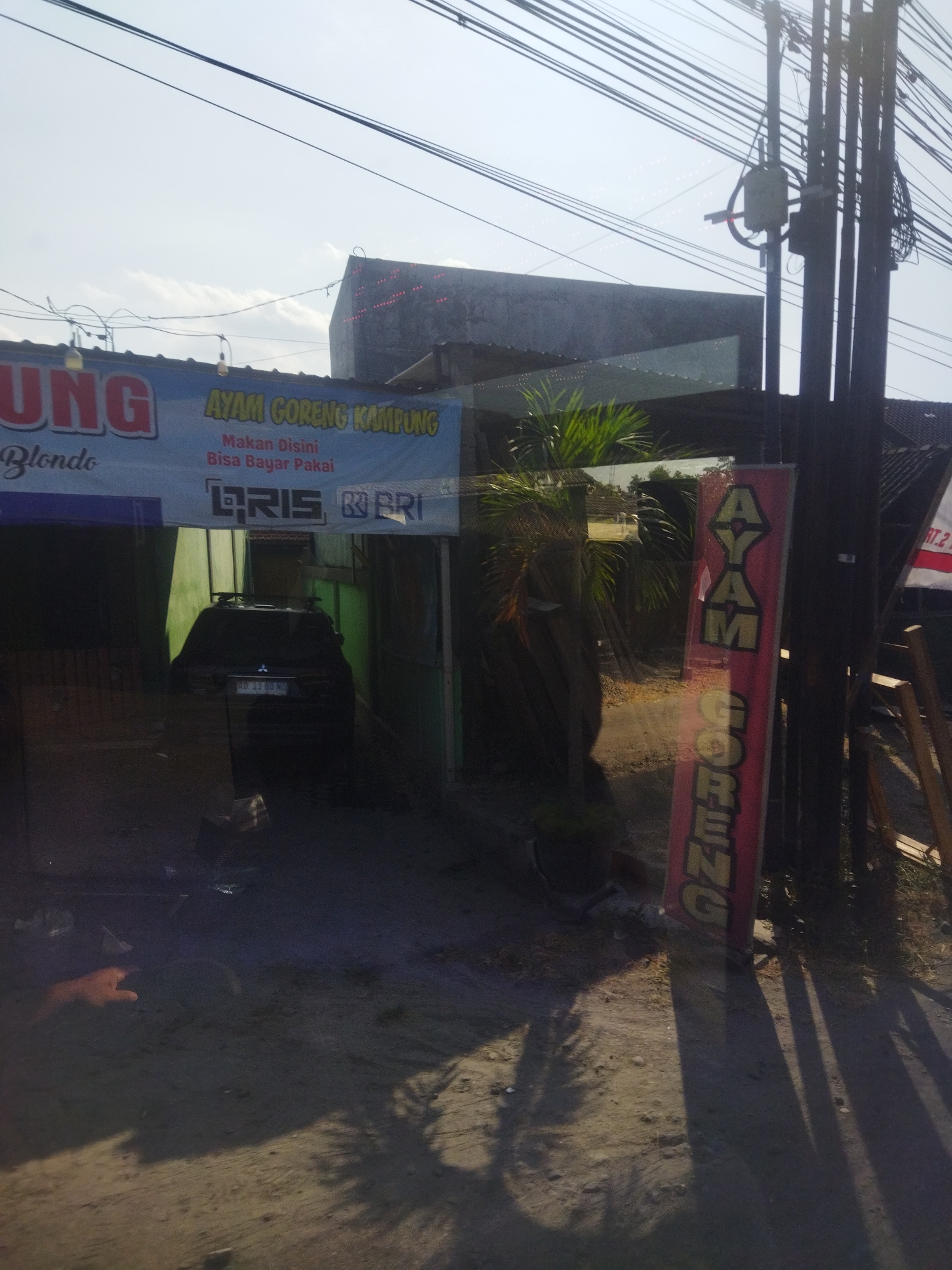 The image depicts a street scene in Nguter, Indonesia, likely near a food stall or restaurant. A banner in the upper left reads "AYAM GORENG KAMPUNG" (Fried Village Chicken) with smaller text below stating "Makan Disini Bisa Bayar Pakai" (Eat Here Can Pay With). To the right of this banner, a vertical red sign with yellow lettering also reads "AYAM GORENG." In the center of the image, a dark-colored SUV is parked. The ground is dirt and gravel, with shadows cast by overhead wires and vegetation. Electrical poles and wires dominate the upper portion of the image, suggesting an urban or semi-urban setting. The lighting suggests it is daytime.
