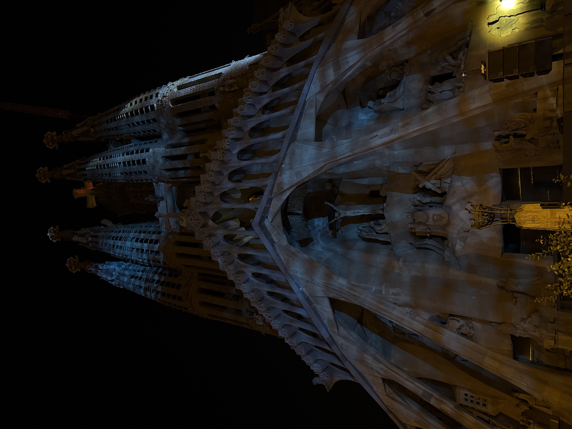 The image shows an architectural structure at night. The stone structure appears to be the Sagrada Familia in Barcelona, Spain. The architecture has multiple spires or towers, along with ornate details and embellishments. The lighting is mostly dark, with artificial illumination on the stone facade. There are no people visible in the scene. The environment consists of the building and dark surrounding space, suggestive of night time.