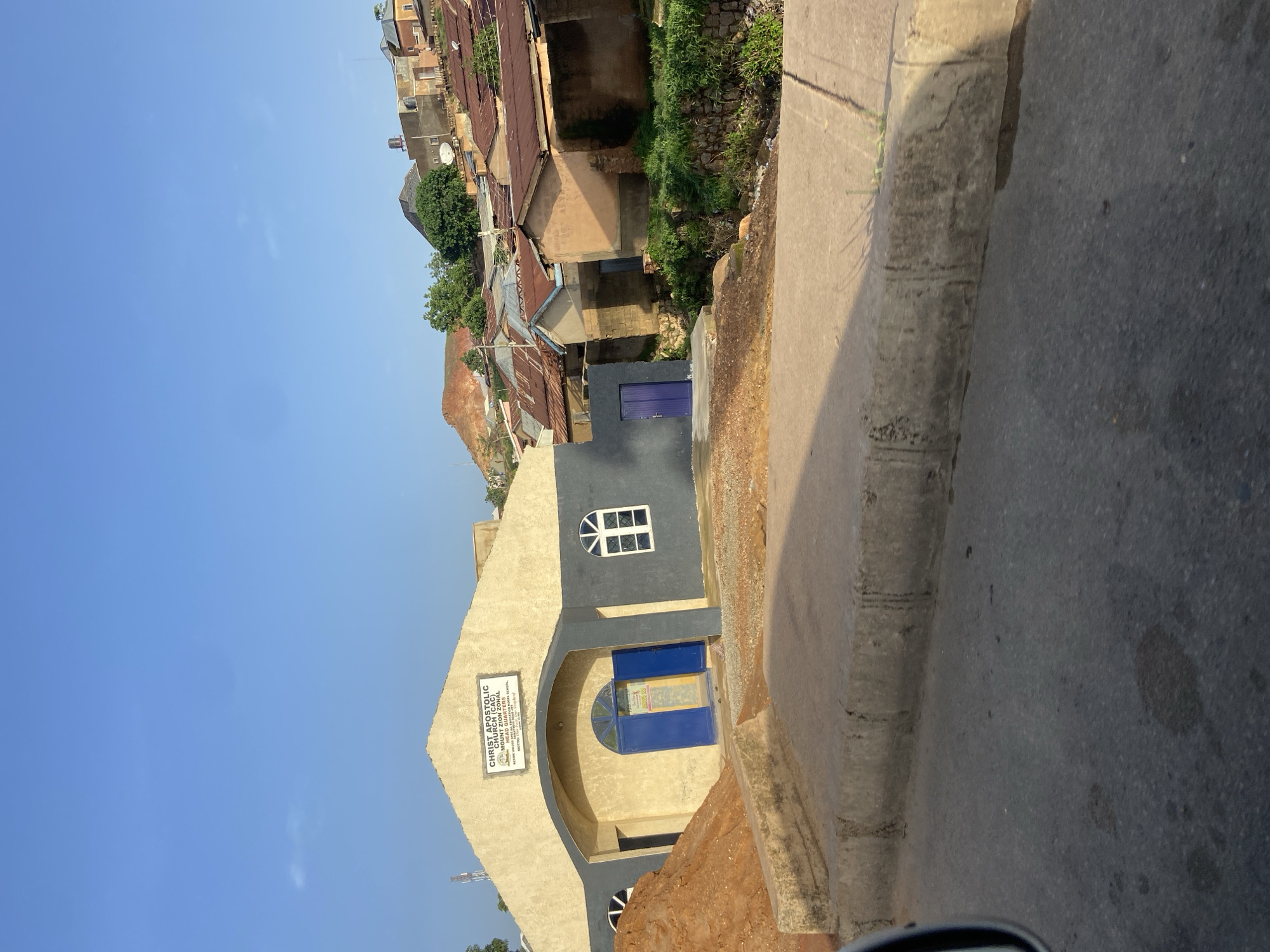 The image shows the exterior of the Christ Apostolic Church (C.A.C) Mount Headquarters in Barakin Lerawi, Nigeria. The building is a light yellow structure with a dark gray facade around the entrance and windows. The main entrance features a large, arched opening with two blue doors and a small sign inside. Above the entrance, a sign reads "CHRIST APOSTOLIC CHURCH (C.A.C) MOUNT HEADQUARTER...". To the right of the entrance, there is a white arched window. Another similar window is visible on the left side of the building, partially obscured by a large pile of reddish-brown soil. To the left of the main structure, a separate dark gray building with a blue door is visible. The church is situated on a slight incline, with several other buildings and rooftops visible in the background, indicative of a densely populated area. A road runs in the foreground, with a concrete curb separating it from the church property. The sky is clear and blue, suggesting a sunny day.