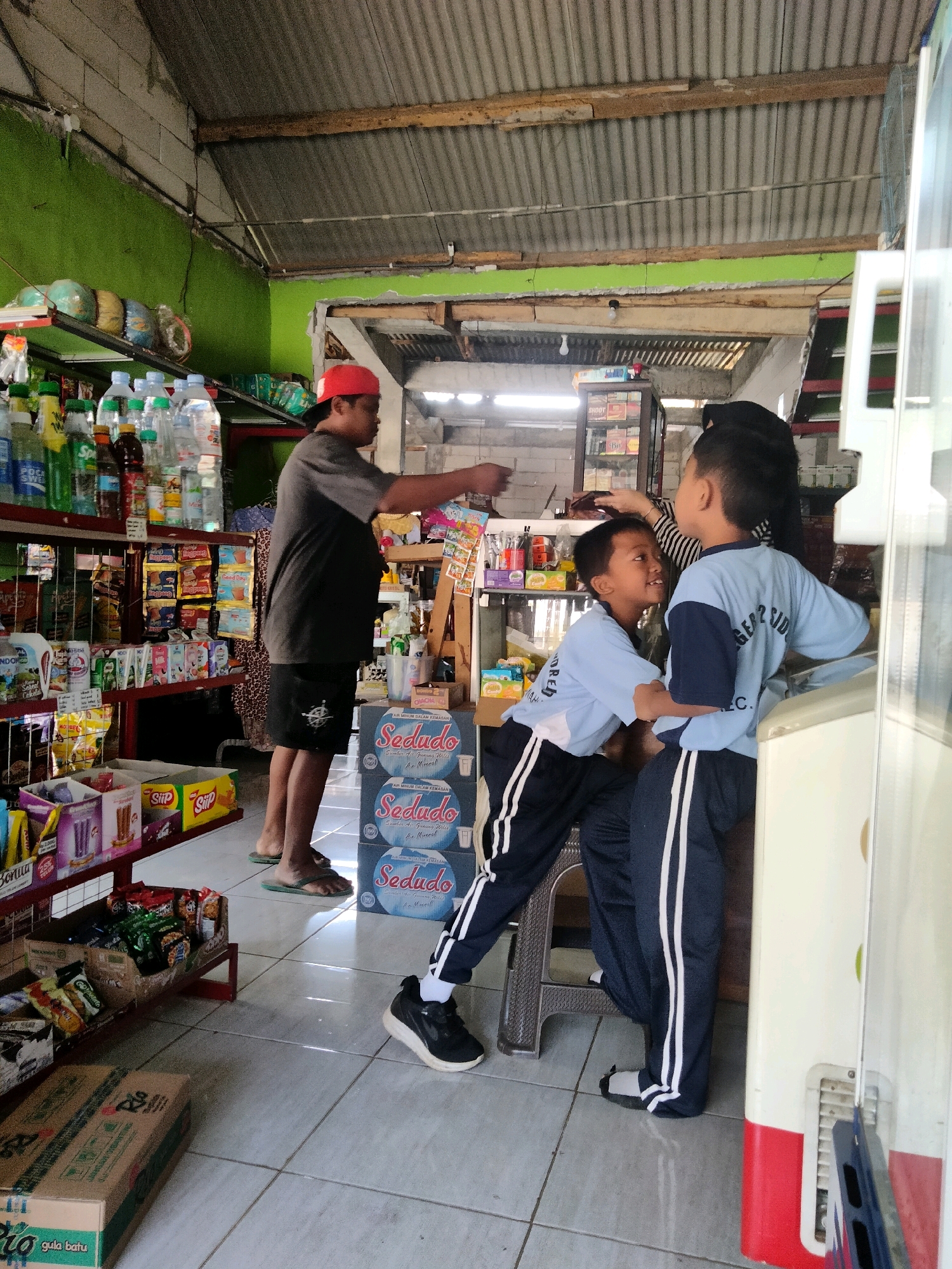 In Sidorejo, Indonesia, within a dimly lit, open-air convenience store, an adult male wearing a red baseball cap and a dark t-shirt is reaching onto a shelf filled with bottled drinks. Two young boys, both in blue and navy school uniforms, are the focal point of the scene. One boy, on the left, leans forward, looking up at the other boy, who is standing and has his hand on the first boy's head. The store's interior is lined with shelves stocked with various food items, snacks, and beverages. A corrugated metal roof is visible overhead. The floor is tiled, and sunlight streams in from the open front, casting shadows. In the foreground, stacked boxes with Indonesian text, likely containing drinks, are visible.