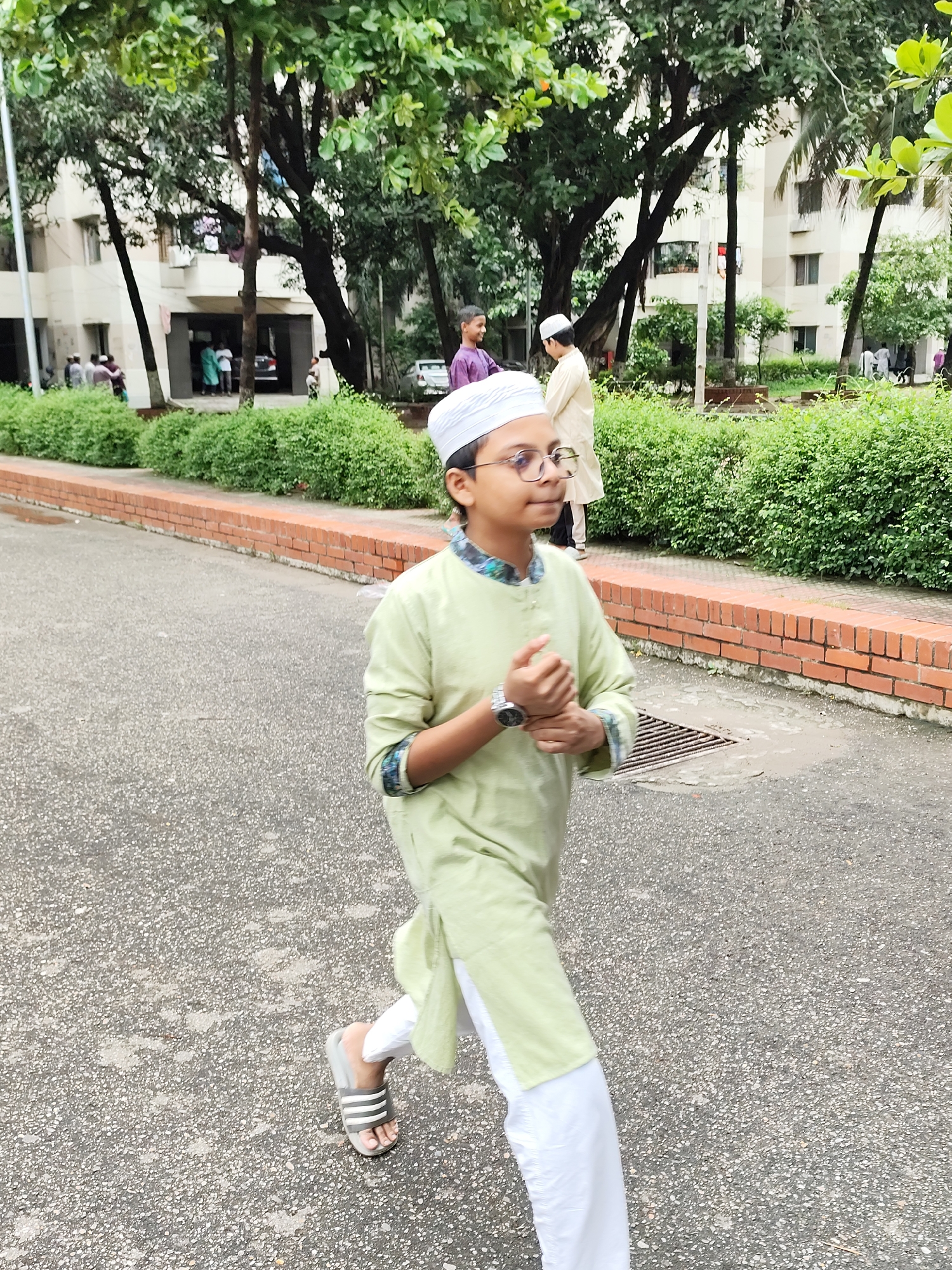 A young boy, wearing a white taqiyah, glasses, a light green kurta, and white pants, walks on an asphalt pathway in what appears to be an urban residential area. He is moving from left to right across the frame and appears to be jogging or walking briskly. He has a wristwatch on his left wrist and is holding his hands clasped together in front of him. The pathway is bordered by a brick wall and a dense green hedge. In the background, there are multi-story buildings with balconies and trees with green foliage. Other individuals, also wearing traditional Islamic attire, are visible in the background near the buildings. The overall scene suggests a warm, possibly humid day. The architecture and vegetation are consistent with an urban setting in Dhaka, Bangladesh.