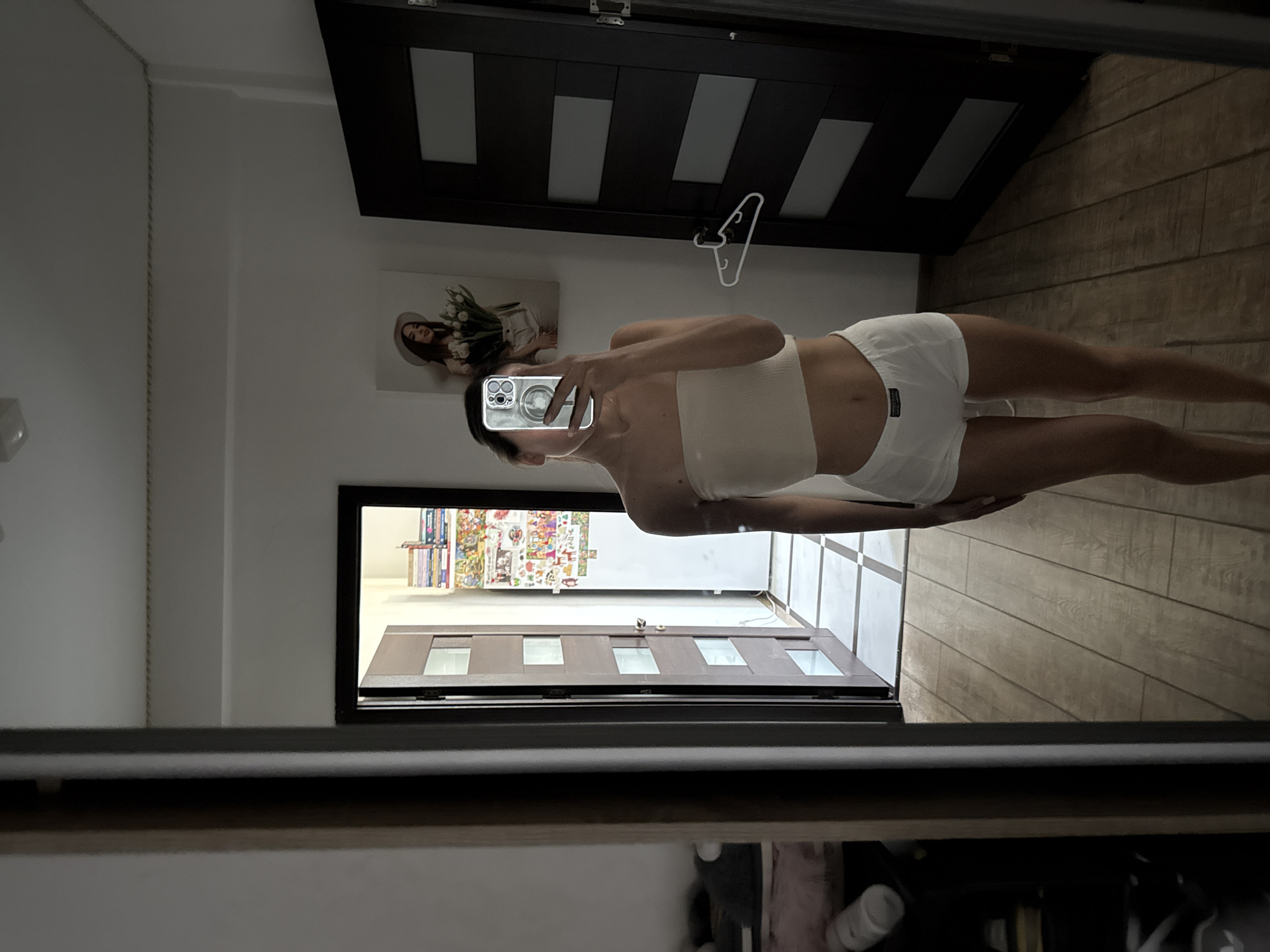 The image shows a young woman taking a mirror selfie in a room. She is wearing a white bandeau top and white shorts. The room appears to be a bedroom or a dressing area, with wooden floors and doors. There is a painting on the wall behind her, and a shelf filled with books and items is visible through an open doorway. The overall lighting suggests it is daytime, with light entering from the open doorway. This scene is not specific to Chișinău, Moldova, or any particular city.