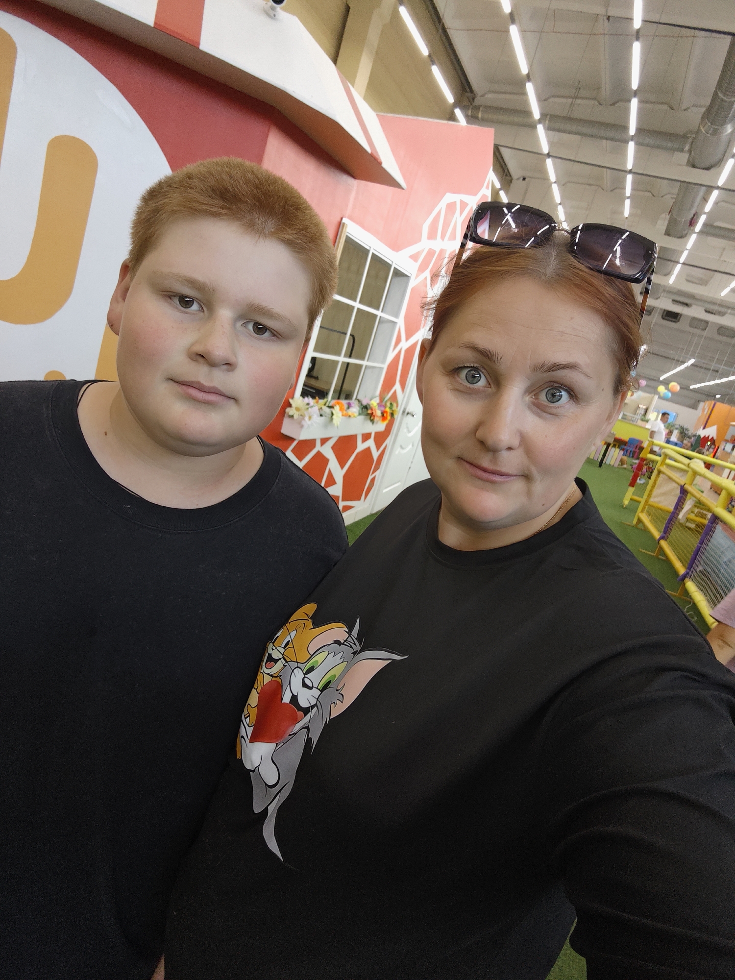 A medium shot shows two people, a young boy and a woman, posing for a selfie indoors. The boy, on the left, has short, reddish-brown hair and is wearing a black t-shirt. He looks directly at the camera with a neutral expression. To his right, the woman has reddish-brown hair pulled back and is wearing a black t-shirt with a graphic of Tom and Jerry holding a heart. Sunglasses are perched on top of her head. Her eyes are wide open, and she has a slight smile.
Behind them, the background suggests an indoor entertainment or play area. There is a colorful, stylized structure that resembles a house with a red and white striped roof and a quirky, slanted design. A window with flower boxes is visible on the structure. The lighting is bright, suggesting daytime, with overhead fluorescent lights. Further back, yellow and purple safety barriers, green turf, and colorful balloons indicate a children's play zone.
There is no visible text in the image. The overall mood appears casual and relaxed, as if the two subjects are enjoying a day out. The location cues point to a public indoor facility, possibly a children's amusement center.