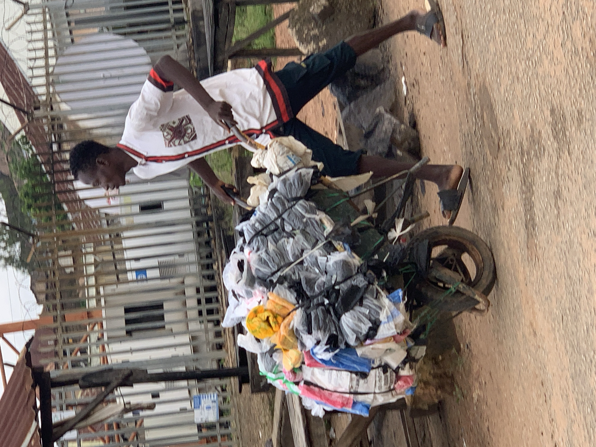 A young Black man is pushing a wheelbarrow filled with plastic bags. He is wearing a white short-sleeved shirt with a red and black trim, and dark green shorts. He has on dark slide sandals. The wheelbarrow is piled high with various plastic bags, some appearing to contain trash or recyclables. The man is in motion, pushing the wheelbarrow forward.
The background shows a fenced area with what appear to be white shipping containers or modular buildings. There are also some metal structures, possibly part of a construction site or industrial area. In the distance, there are buildings and some vegetation. The ground is a reddish-brown, dusty surface, suggesting a somewhat arid environment. The scene appears to be outdoors, possibly in an urban or peri-urban area. The overall impression is one of everyday work and activity.