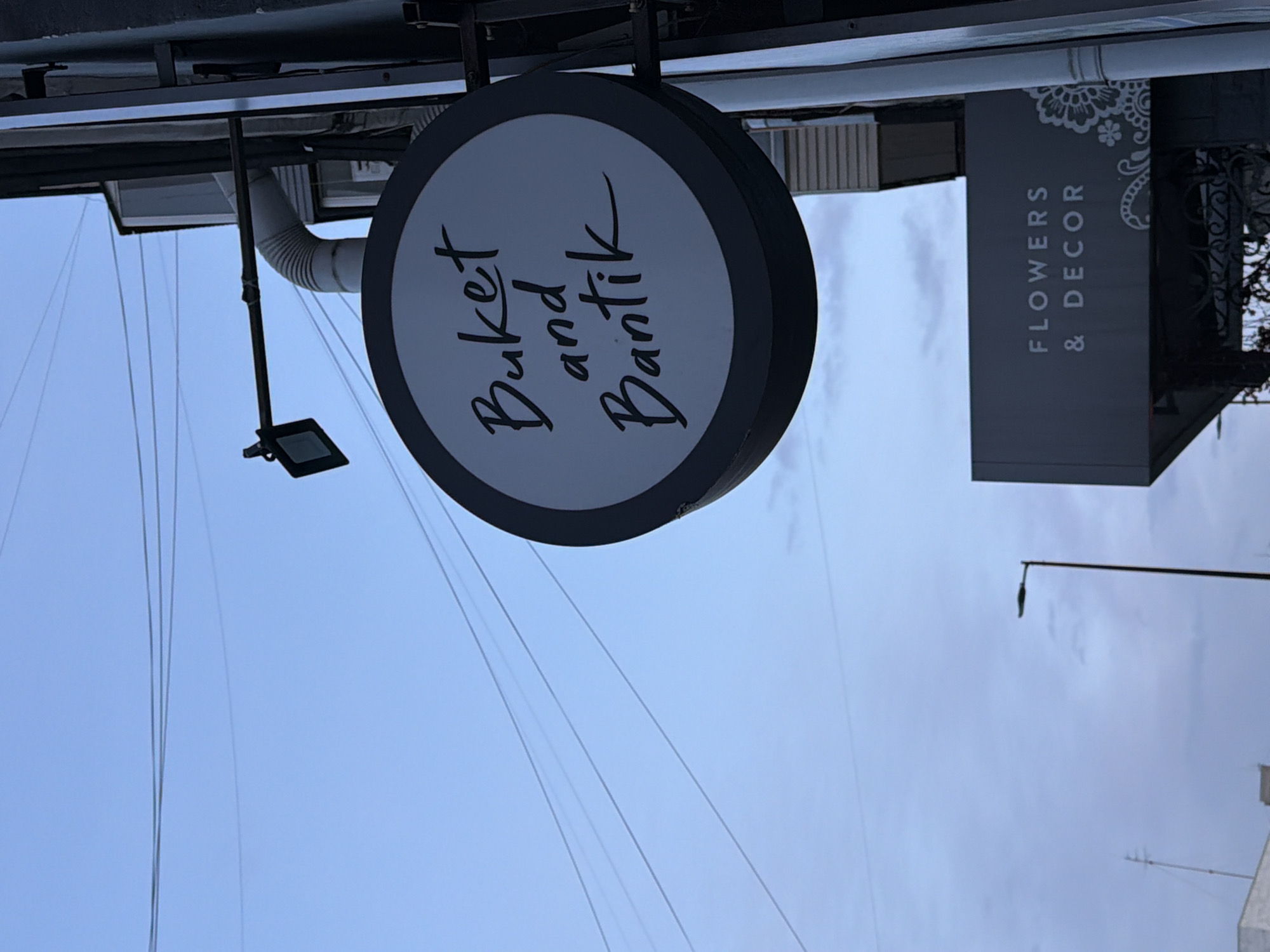 Here's a description of the image:
The image shows outdoor signage for a business, with a partial view of the sky in the background. There are two primary signs visible:
1. **Round Sign:** A circular sign with a dark gray rim and a light gray/white center. The words "Buket and Bantik" are written in a cursive black font in the center.
2. **Rectangular Sign:** A vertical rectangular sign to the right of the round sign. The words "Flowers & Decor" are vertically stacked in a white font against a dark gray background. There is a decorative floral design at the top of the sign.
The signage is attached to a building or structure, with supporting metal bars and a lighting fixture visible above the round sign. The image is rotated, the direction is horizontal.
Based on the Cyrillic transliteration in the signage ("Buket and Bantik"), it is likely that the business is located in a region where Russian is spoken. Given the additional information that the location is in Krasnoyarsk, Russia, it can be inferred that the shop providing floral and decorative services, and the image itself is situated there.