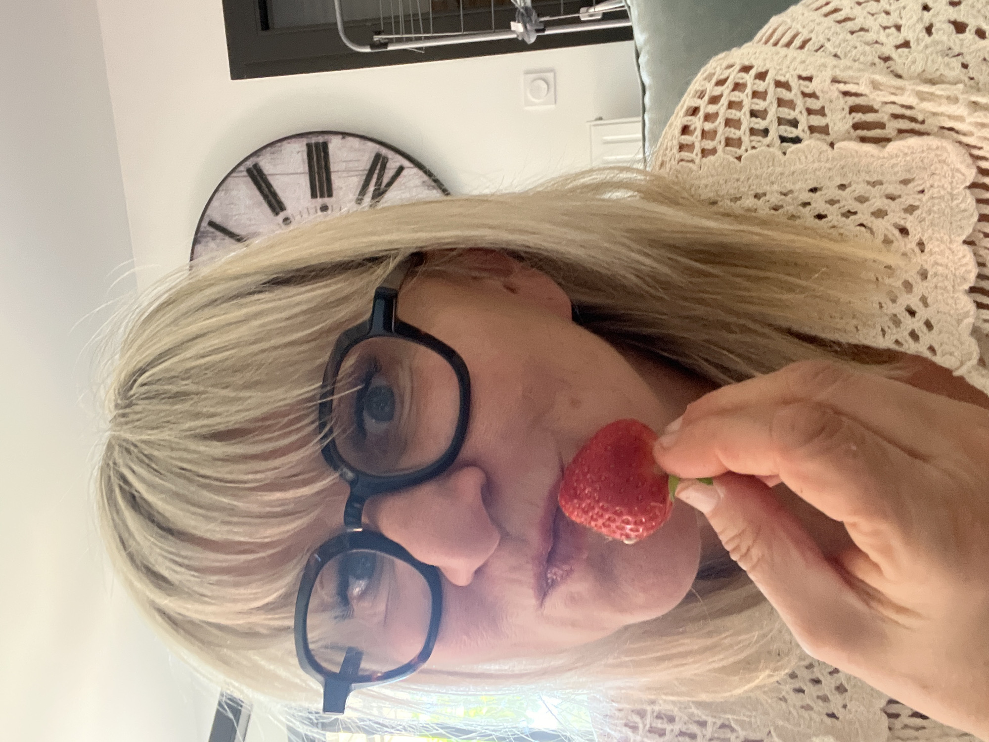 A woman with blonde hair and black glasses holds a strawberry in front of her mouth. She is wearing a crocheted shawl. In the background is a wall clock and part of a metal structure. The location is identified as Chambœuf, France.