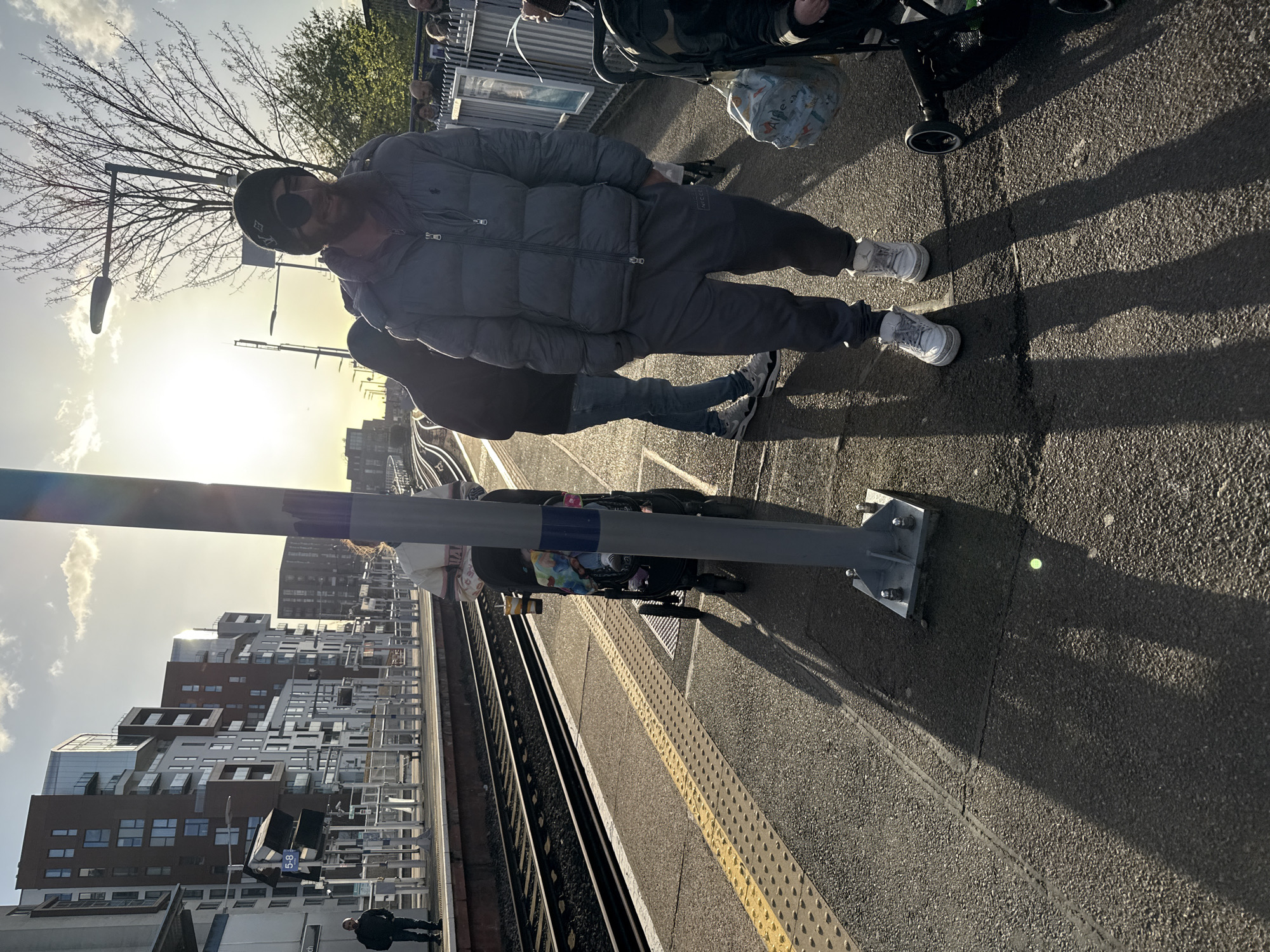 An individual stands on a train platform next to a stroller. The platform is demarcated by a textured yellow line adjacent to the train tracks. Across the tracks are buildings, and a light pole is visible in the foreground. The man is wearing a dark hat, sunglasses, a gray puffer jacket, and matching sweatpants and shoes. Other people are in the background. The location is likely in a modern area of London, United Kingdom, given the style of architecture.