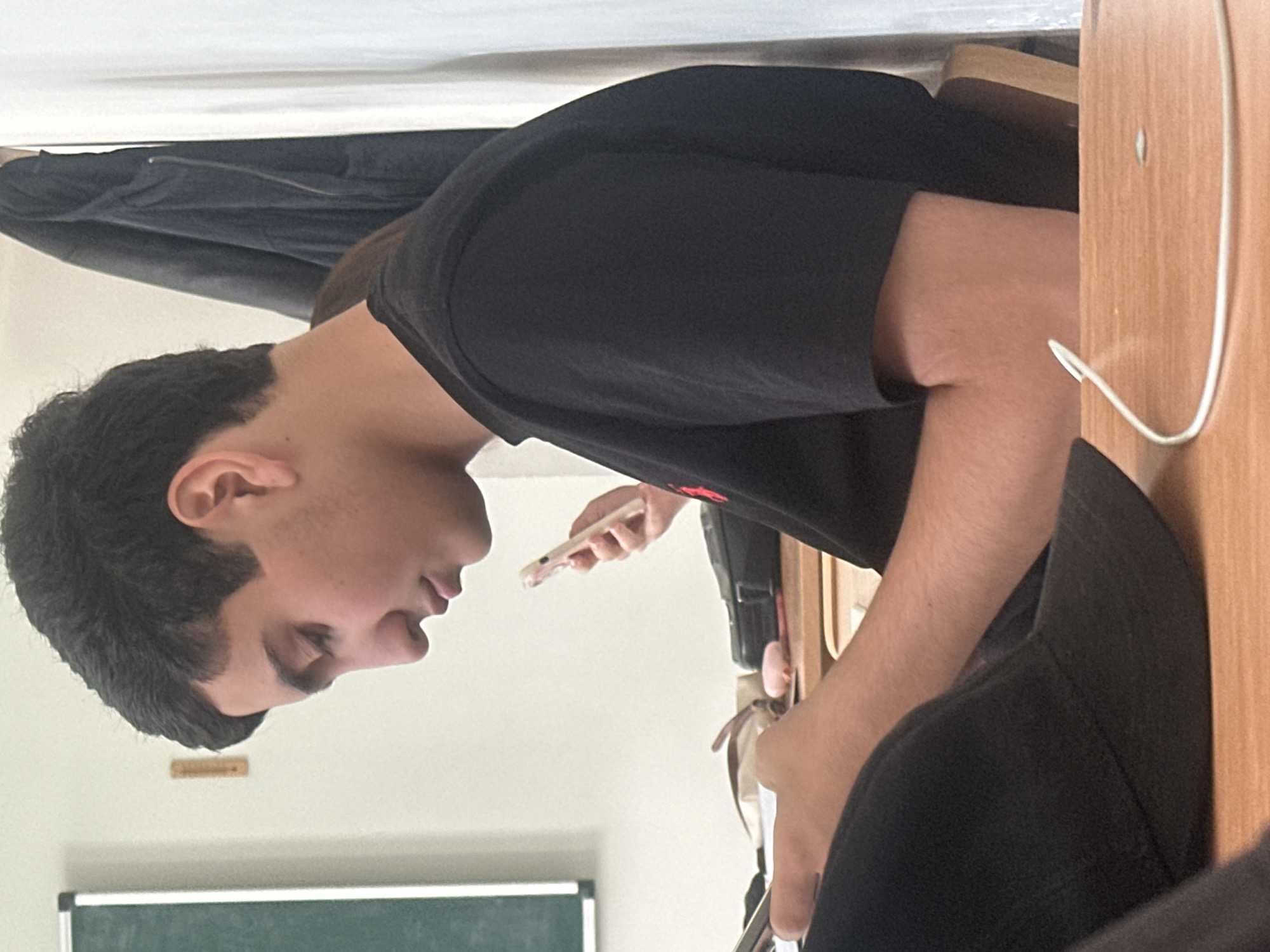 A medium shot captures a young male individual seated at a wooden desk in what appears to be a classroom in Yerevan, Armenia. The subject, positioned in profile facing left, has dark, short hair and light skin, and is wearing a black short-sleeved t-shirt. His gaze is directed downwards, suggesting focus on something below the frame, possibly a smartphone which is partially obscured by his right arm. Another person's arm, holding a light-colored smartphone, is visible in the background, closer to the subject's head.
The setting includes a light-colored wall in the background, with a dark green chalkboard framed in white visible at the bottom left. A small wooden object, possibly a ruler, is mounted on the wall. A dark jacket or garment hangs behind the main subject, and a black bag or case is on the desk near his right arm. A white cable runs across the wooden desk surface in the foreground, suggesting a charging cable or headphone wire. The overall atmosphere is quiet, characteristic of a study or classroom environment. The subject's expression is neutral and attentive. No visible text is discernible in the image. The time of day is indeterminate, but the indoor lighting suggests daylight.