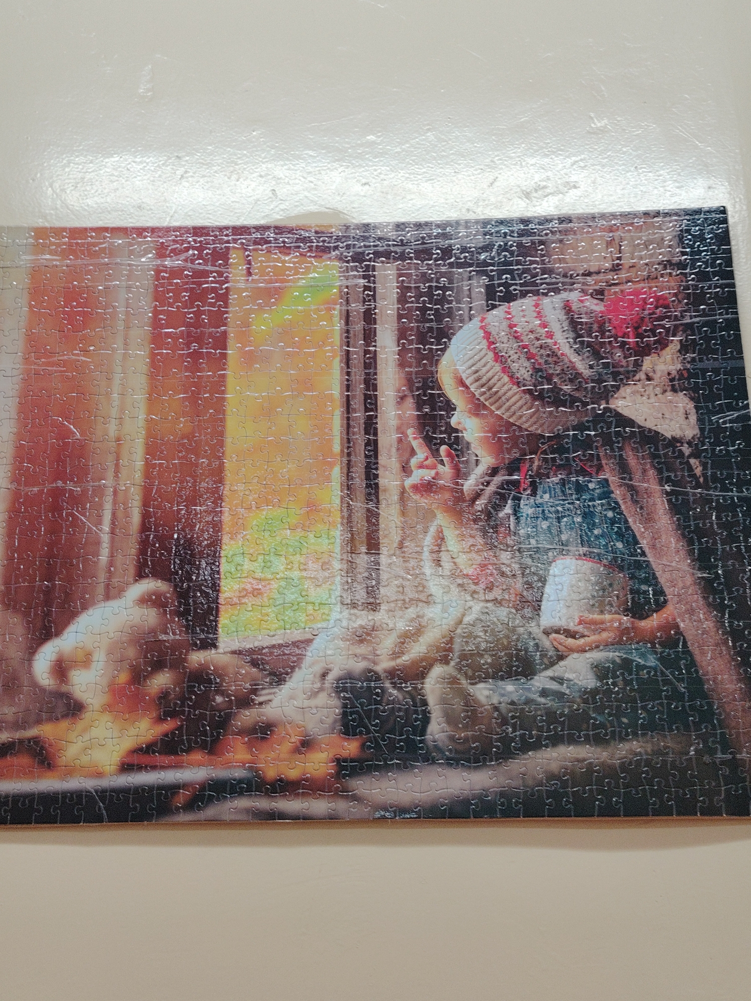 A completed jigsaw puzzle depicts a child in profile sitting by a window. The child wears a striped beanie and overalls and holds a white mug. Outside the window, there are blurred colors suggesting trees, possibly an autumn scene. A stuffed animal sits on the windowsill to the left. The location is not discernible from the image, and there's no indication this photo was taken in Sarapul, Russia.
