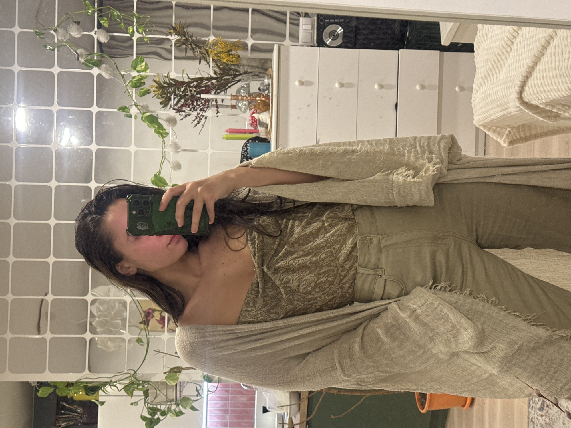A person is taking a selfie in a bathroom. The person has long, dark hair and is wearing a patterned top, jeans, and a light-colored shawl. The bathroom has tiled walls and a cabinet. Plants are visible in the reflection. There is no evidence to suggest this image was taken in Krasnoyarsk, Russia.