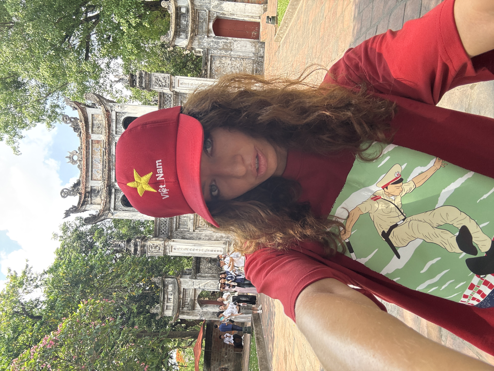 A selfie shows a woman wearing a red hat with a yellow star and the words "Việt Nam" embroidered on it. She is also wearing a red shirt with a graphic print of a police officer on it. Behind her is an ornate stone gate and a group of people. The location is likely a historical or cultural site in Hà Nội, Vietnam, given the architecture.