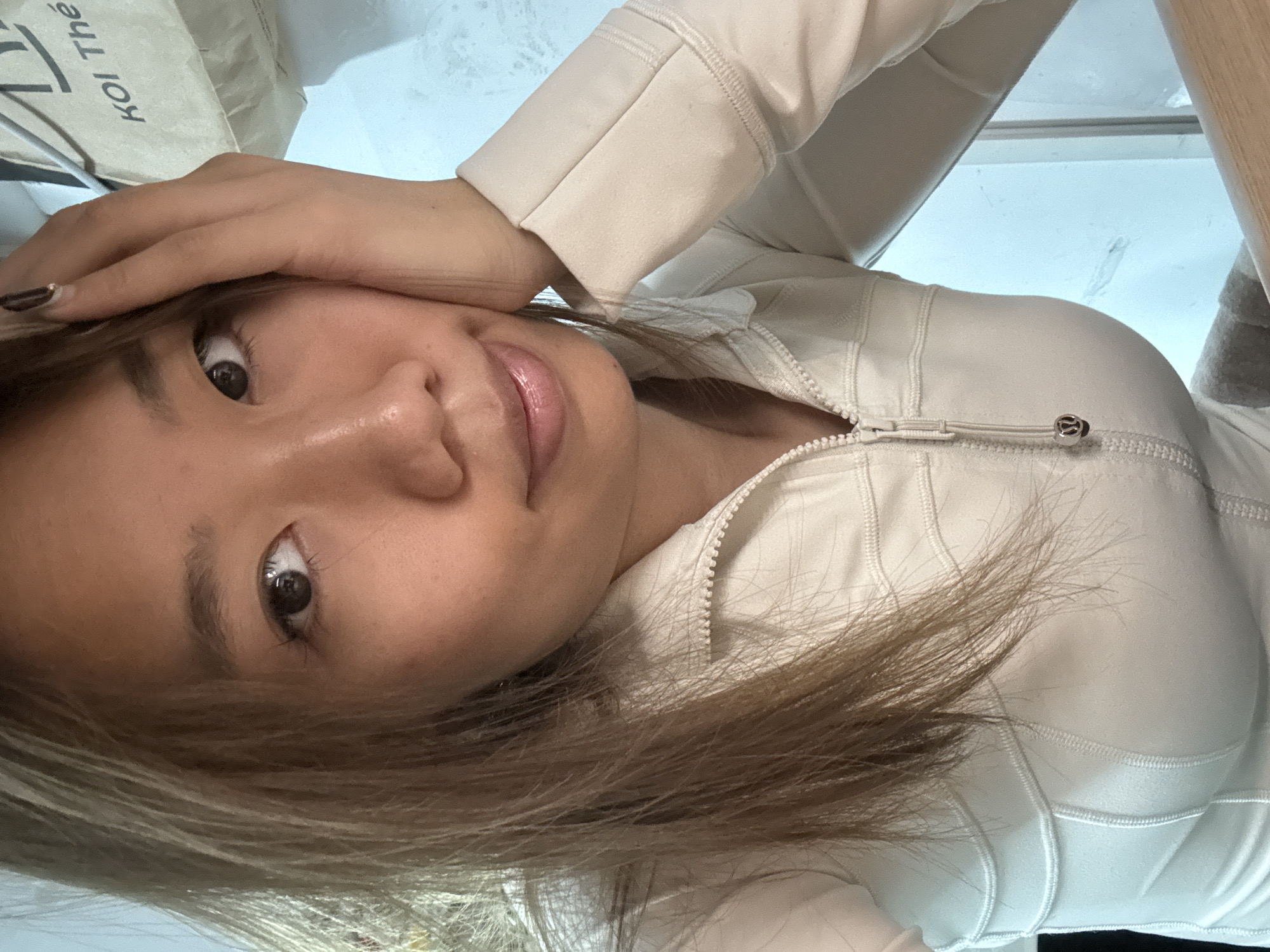 A woman is pictured indoors, likely in a residential setting. She has light brown hair with blonde highlights, and brown eyes. She is wearing a light-colored zip-up jacket. Her left hand is resting on her forehead. A paper bag with the name "Koi Thé" printed on it is visible in the upper left corner of the frame. The background shows a blurred mix of light and dark colors, suggesting a window or other indoor elements. It is impossible to determine if the image was taken in Nan'an District, China, without further information.