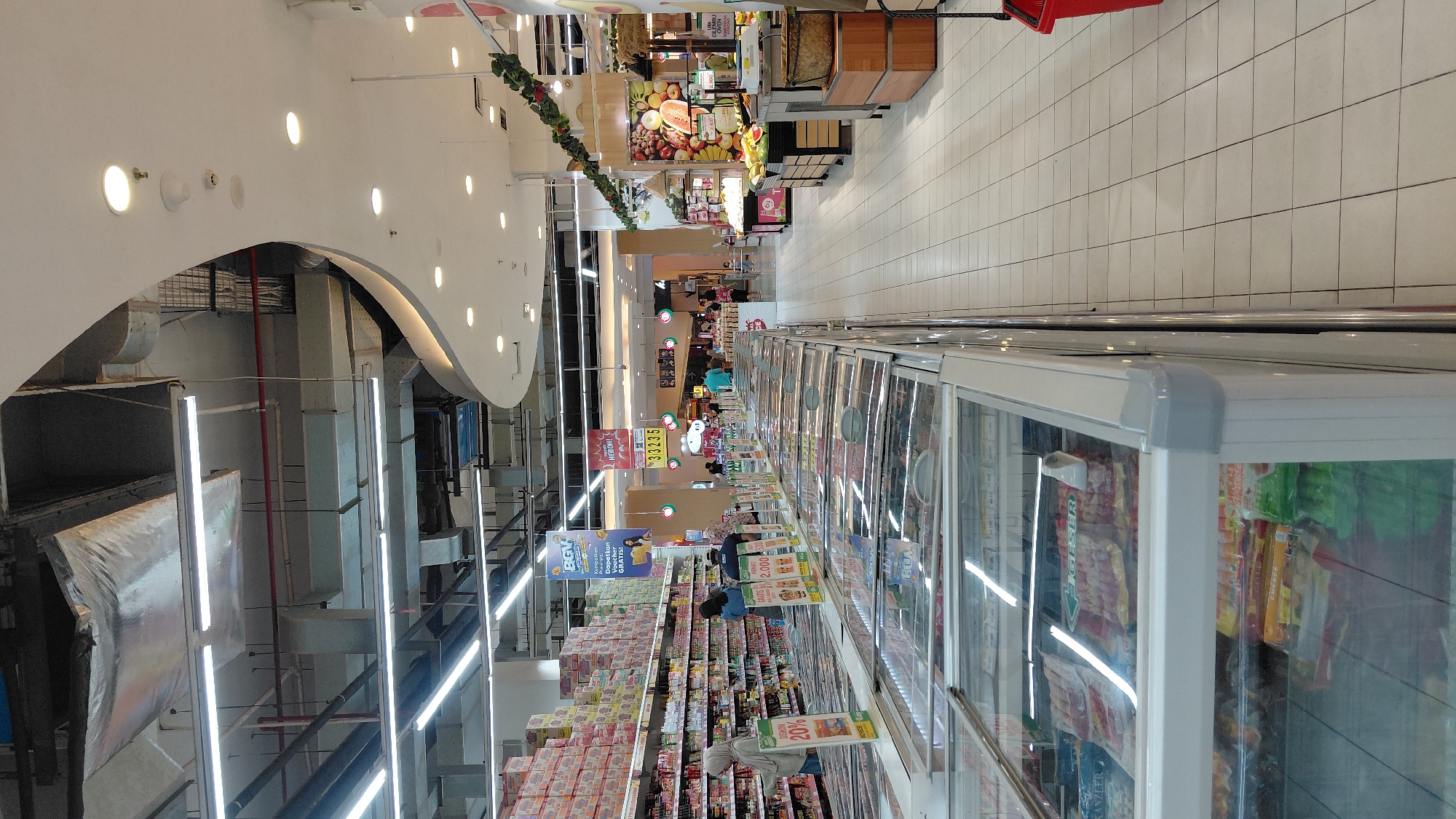 The image depicts the interior of a brightly lit supermarket, likely in Purwokerto, Indonesia, given the signage and product packaging visible. The perspective is from the aisle, looking down a long row of refrigerated display cases filled with frozen goods such as sausages and other packaged food items. The aisle flooring is tiled in a light color, creating a clean and organized appearance.
To the left, towering shelves are stocked with various boxed products, primarily food items like instant noodles. A woman wearing a hijab is browsing the shelves. Further down the aisle, several other shoppers are visible, some interacting with displays or walking through the store. Several promotional signs are displayed throughout the supermarket, advertising discounts and special offers. For example, one sign prominently displays "DISCOUNT 20%". Another sign reads "HEBOH!!!" with a price of "33235".
The ceiling features a complex network of industrial lighting fixtures, air conditioning ducts, and electrical conduits, with a distinctive curved, white architectural element adding a modern touch to the space. On the right side of the image, a section of the store dedicated to fresh produce is visible, with colorful fruits and vegetables displayed. The overall impression is of a busy, well-stocked, and modern retail environment.