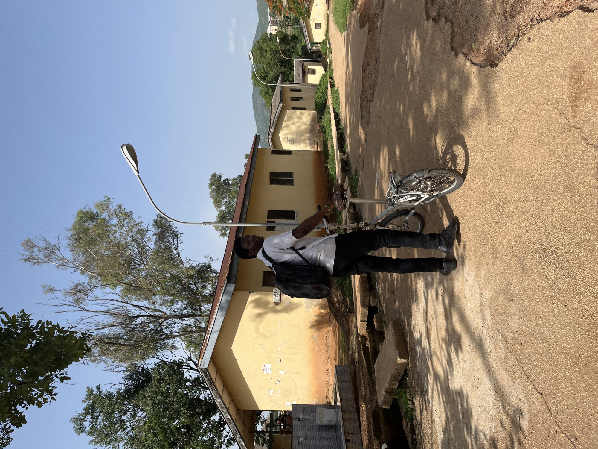 In Jos, Nigeria, a young Black male wearing a white short-sleeved shirt, black trousers, and a black backpack stands next to a silver bicycle on a dirt and asphalt road. He is looking over his left shoulder towards the camera. Behind him, a single-story yellow building with a rusty metal roof is visible, alongside a street lamp. The building has several windows and is adorned with some signs and peeling paint. The environment suggests a somewhat rural or institutional setting, possibly a school or residential compound, with trees and vegetation in the background and a clear blue sky overhead. The ground is uneven, with patches of gravel and some concrete slabs.