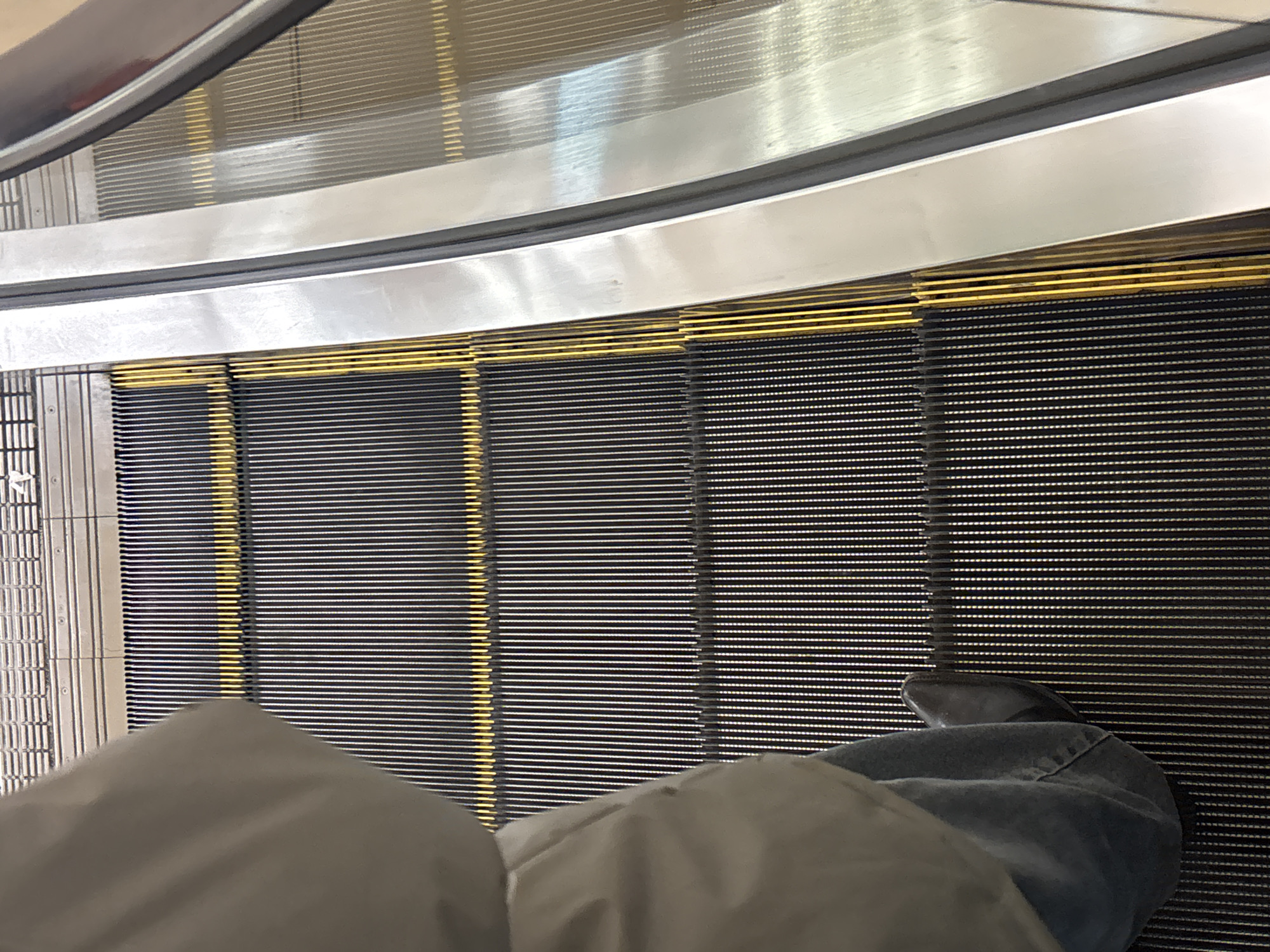The image shows a person riding an escalator. The person is wearing grey pants and a shoe. The escalator is made of black and gold metal. The handrail is silver. The location is unknown and not identifiable as Perm, Russia.
