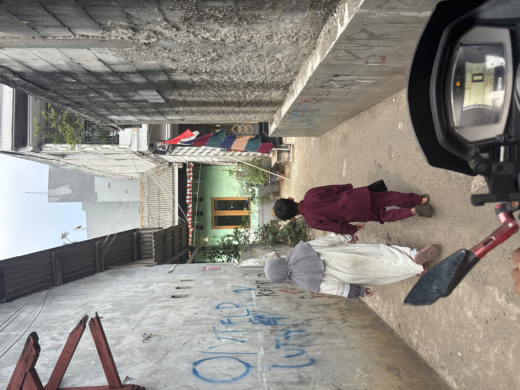 Two children, a girl wearing a grey hijab and a white patterned dress, and a boy in a maroon outfit, walk hand-in-hand down a narrow, paved alleyway. The alley is bordered by textured concrete walls on the right and a rough concrete wall with blue graffiti on the left. Overhead, a corrugated metal roof extends from buildings on the left. On the right, a multi-story building is visible with a balcony on the second floor and a decorative flag hanging from a pole. A green house with a wooden-framed window is visible at the end of the alley on the left, adorned with potted plants. The foreground of the image shows the dashboard of a motorcycle, with a digital display showing "0" and other indicators. The scene appears to be in a residential area in Bekasi, Indonesia.