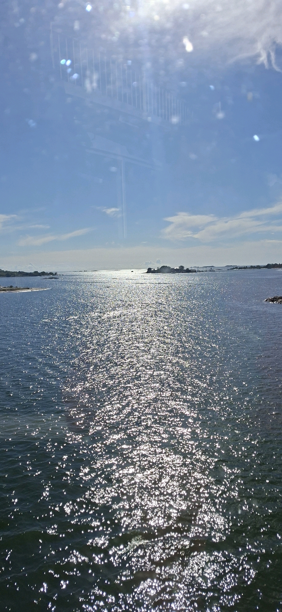 A vertical, full shot captures a sun-drenched expanse of the Baltic Sea, with a sunlit path of sparkles stretching towards the horizon. The sky above is a clear blue, punctuated by sparse, wispy clouds. In the distance, a chain of small, tree-covered islands forms a dark silhouette against the bright water. On the right side of the frame, a large rock protrudes from the sea. The overall impression is one of a serene and sunny day in a coastal environment, possibly near Helsinki, Finland. The water appears calm, with gentle ripples catching the sunlight. The reflections in the water suggest a clear, bright day.