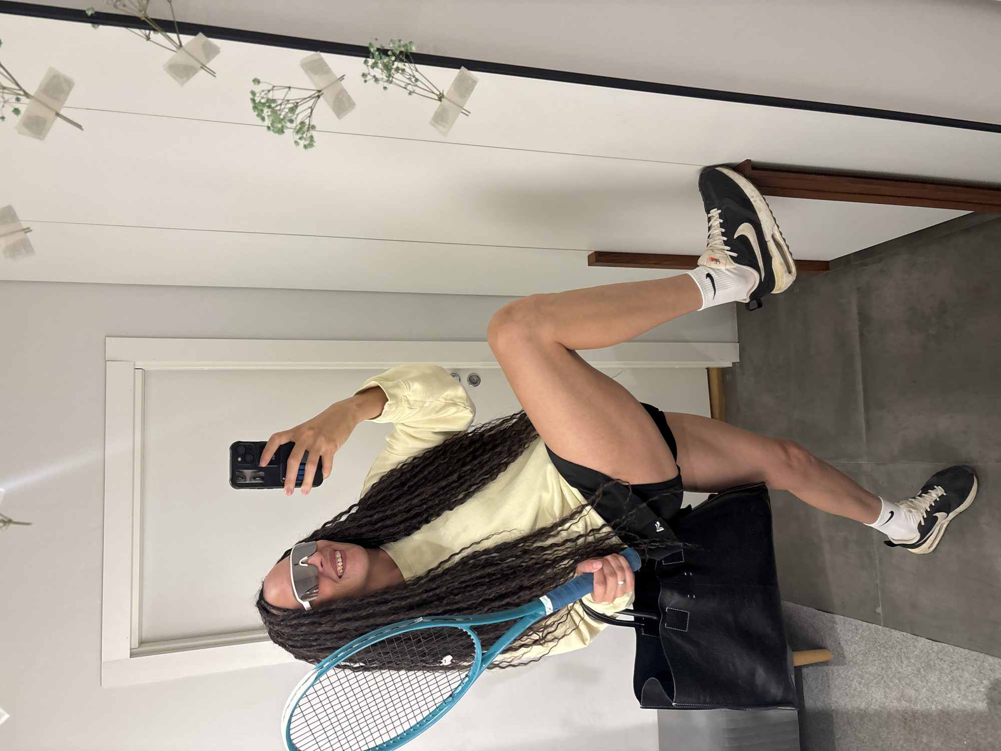 A young woman is captured in a mirror selfie, posing with a tennis racket in what appears to be an indoor setting, possibly in Chaikovsky, Russia. The image shows her with long, dark, curly hair, wearing white cat-eye sunglasses, a light yellow long-sleeve top, and black athletic shorts. She is smiling broadly, revealing her teeth, and holds a black smartphone in her right hand, capturing the reflection. In her left hand, she holds a blue/turquoise tennis racket.
Her legs are prominently featured, one bent at the knee and the other extended. She wears white crew socks with a visible black Nike swoosh logo and black sneakers with white soles and laces. Her left leg rests on a large black tote bag with white stitching, which is placed on a light grey floor. The background of the mirror reflection shows a white wall decorated with delicate sprigs of greenery (possibly baby's breath) taped with white adhesive to thin strings, creating a minimalist, decorative touch. The overall impression is playful and active, suggesting readiness for or participation in a sport like tennis. The indoor lighting does not indicate a specific time of day.
