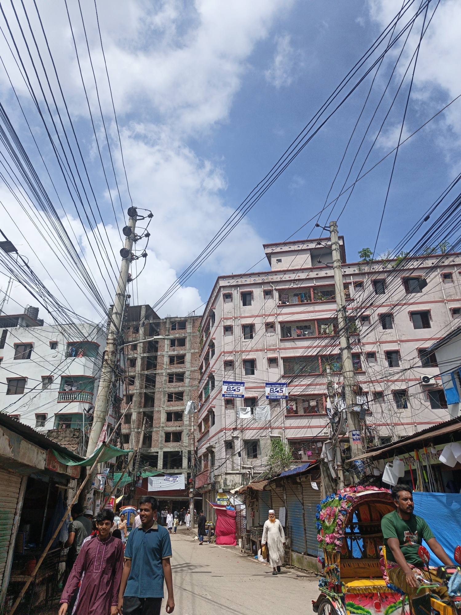 The image depicts a narrow street in Dhaka, Bangladesh, characterized by dense overhead wiring and multi-story buildings. Two young men are walking towards the camera, the one on the left wearing a maroon kurta and the one on the right in a blue polo shirt. To their right, a brightly decorated cycle rickshaw is parked, with its driver seated and looking to the left. Further down the street, other pedestrians can be seen, including a man in a white thobe. The buildings on either side of the street show varying states of construction and occupancy, with some displaying visible laundry hanging from windows. The sky above is blue with scattered clouds, and a prominent utility pole with a complex network of wires dominates the central part of the frame. The overall impression is one of a bustling urban environment with a dense infrastructure.