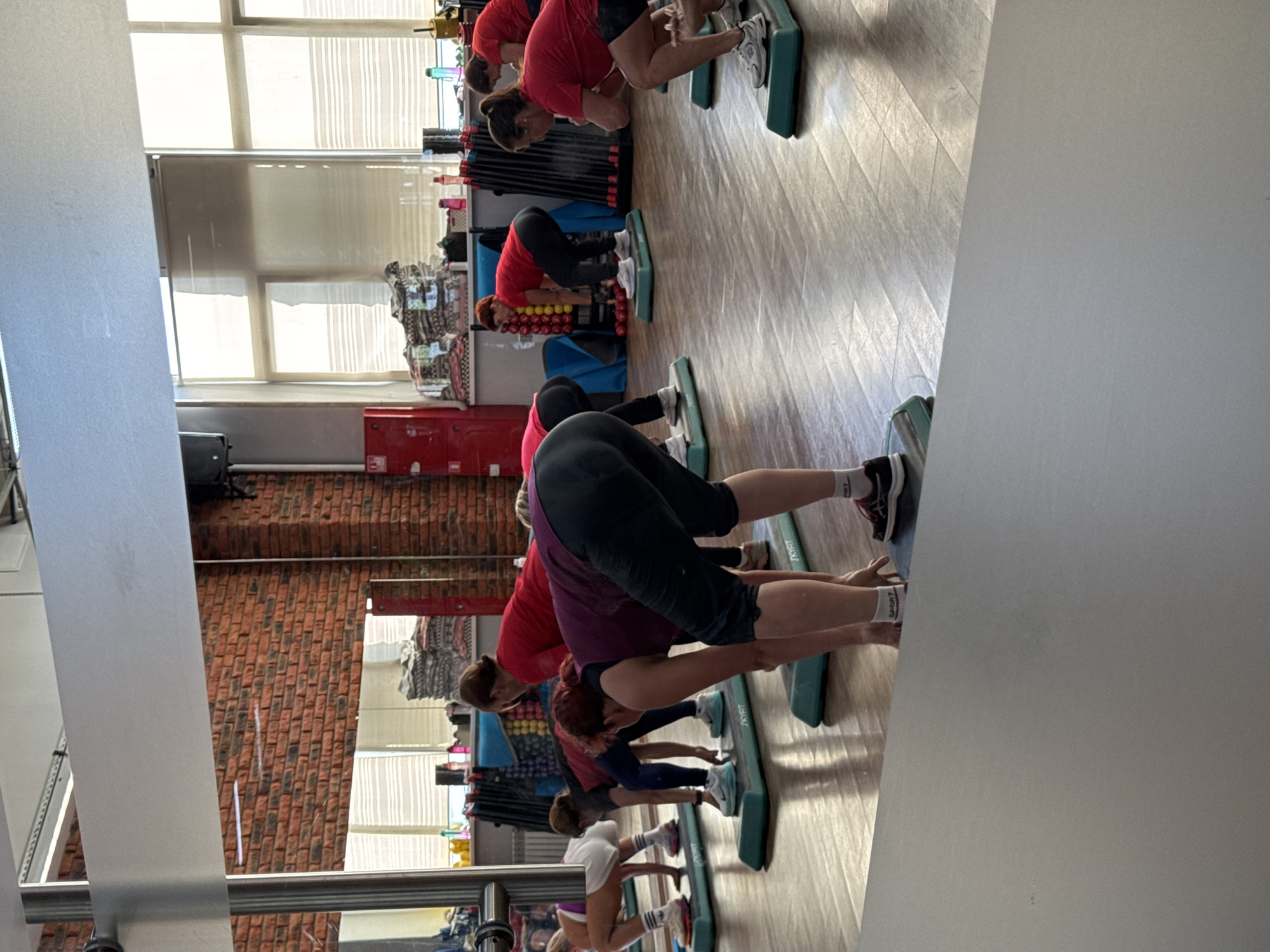 This image shows a group of women participating in a fitness class at a gym. The women are of various ages and are dressed in athletic wear. They are positioned on aerobic steppers and are engaged in an exercise that involves bending at the waist with their hands near their feet. The gym appears to be well-lit with large windows and mirrors. In the background, there are exercise equipment, weights, and storage racks. The floor is made of wood. The overall atmosphere suggests a structured fitness environment.