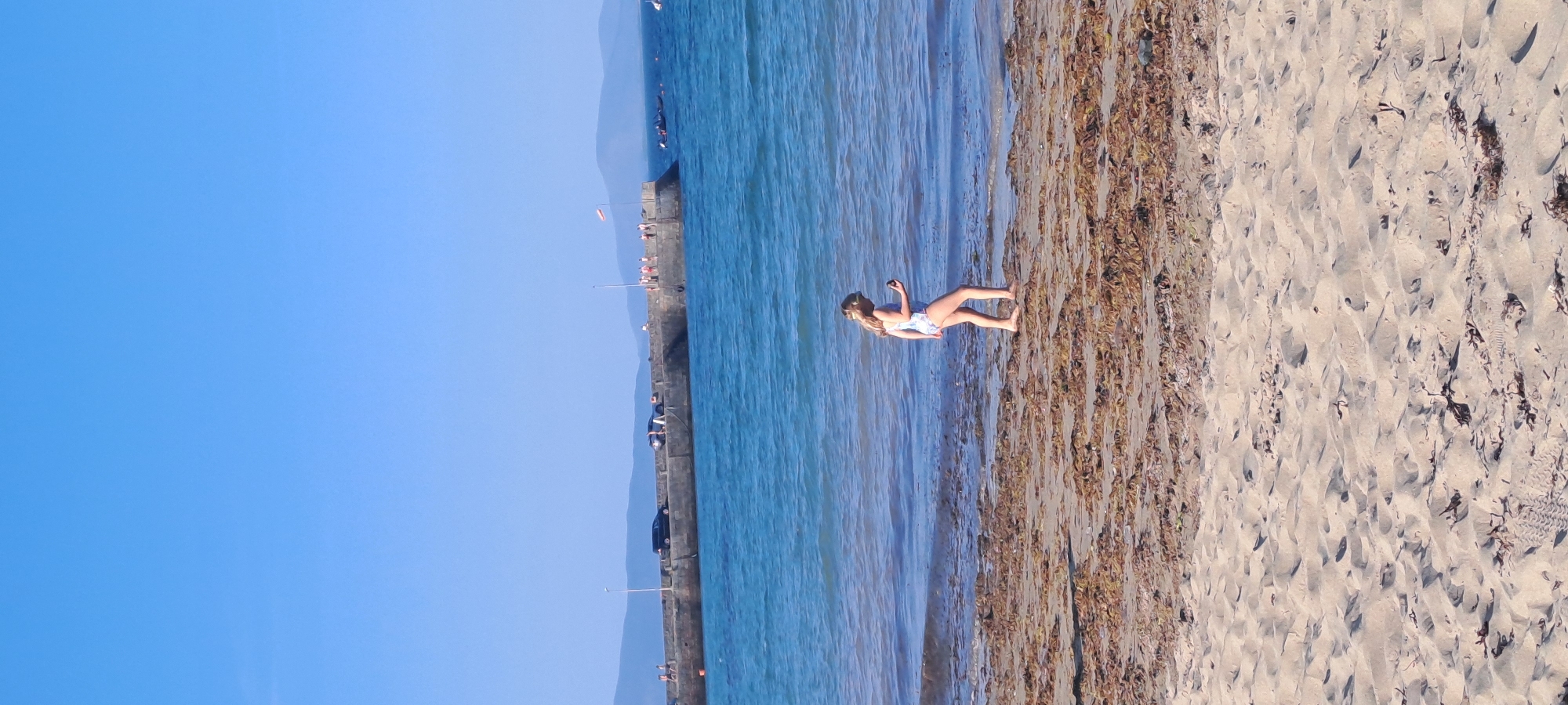 This is a vertical, eye-level, outdoor, medium shot photograph taken on a sunny day at a beach in an unknown location in Ireland. The foreground is a sandy beach littered with brown seaweed. In the middle ground, a young girl with blonde hair wearing a blue and white swimsuit and a headband walks on the wet sand near the water's edge. She is holding a dark object in her right hand and appears to be looking down at it. The water is blue and calm with small waves lapping at the shore. In the background, a long, concrete pier juts out into the sea. Several people are visible on the pier, as well as two cars. Further in the distance, mountains are visible on the horizon under a clear blue sky. The scene appears peaceful and recreational.