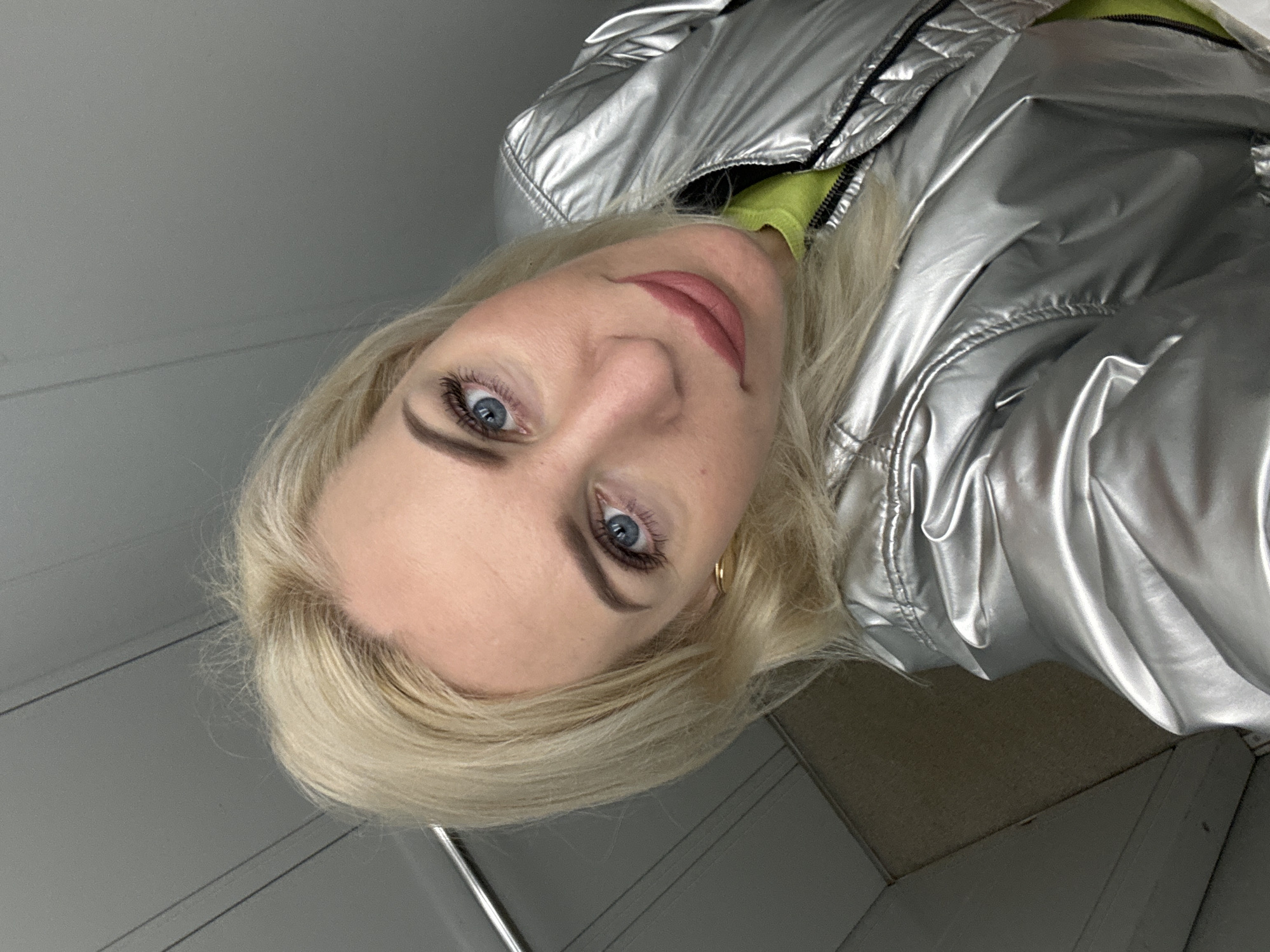 A close-up, tilted portrait shows a fair-skinned woman with blonde hair and blue eyes. She is wearing a shiny silver jacket with black trim and a neon yellow garment underneath. She is wearing gold hoop earrings and visible makeup. The background appears to be a room with gray and white paneling. The location is Unknown, Unknown.