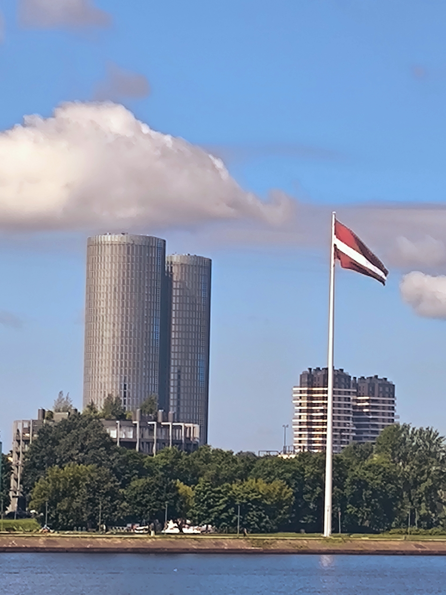 The image shows a scenic view of the Daugava River in Riga, Latvia. In the background, there are several modern buildings, including two prominent cylindrical towers and two rectangular high-rises. The Latvian flag is prominently displayed on a flagpole in the foreground, fluttering in the breeze. Lush green trees line the riverbank, creating a vibrant contrast with the urban architecture. The sky is a clear blue with some scattered clouds, suggesting a pleasant day. The river in the foreground is calm and reflects the sky and surrounding scenery.