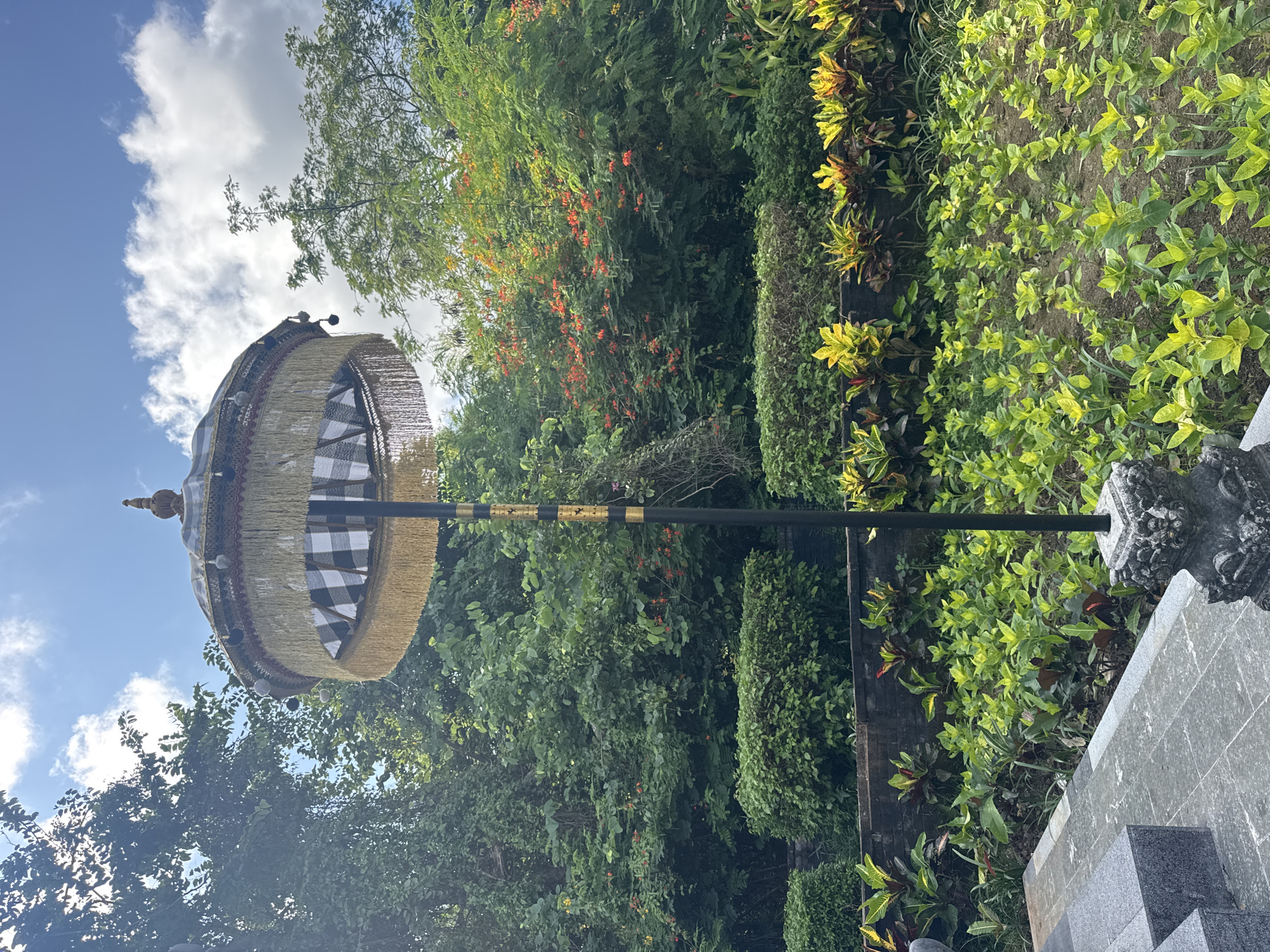 A medium shot shows a Balinese ceremonial umbrella positioned in a lush, green environment in Kuta Selatan, Indonesia. The umbrella, known as Tedung, is elevated on a tall black pole with gold accents. The umbrella's canopy features a black and white checkered pattern, adorned with layers of tassels and decorative spheres. It faces towards the upper left against a backdrop of a cloudy sky. Dense foliage, including trees and various shrubs, forms the background. Some plants show vibrant shades of green and yellow. A stone structure is visible on the right side of the frame, partially obscured by the vegetation.