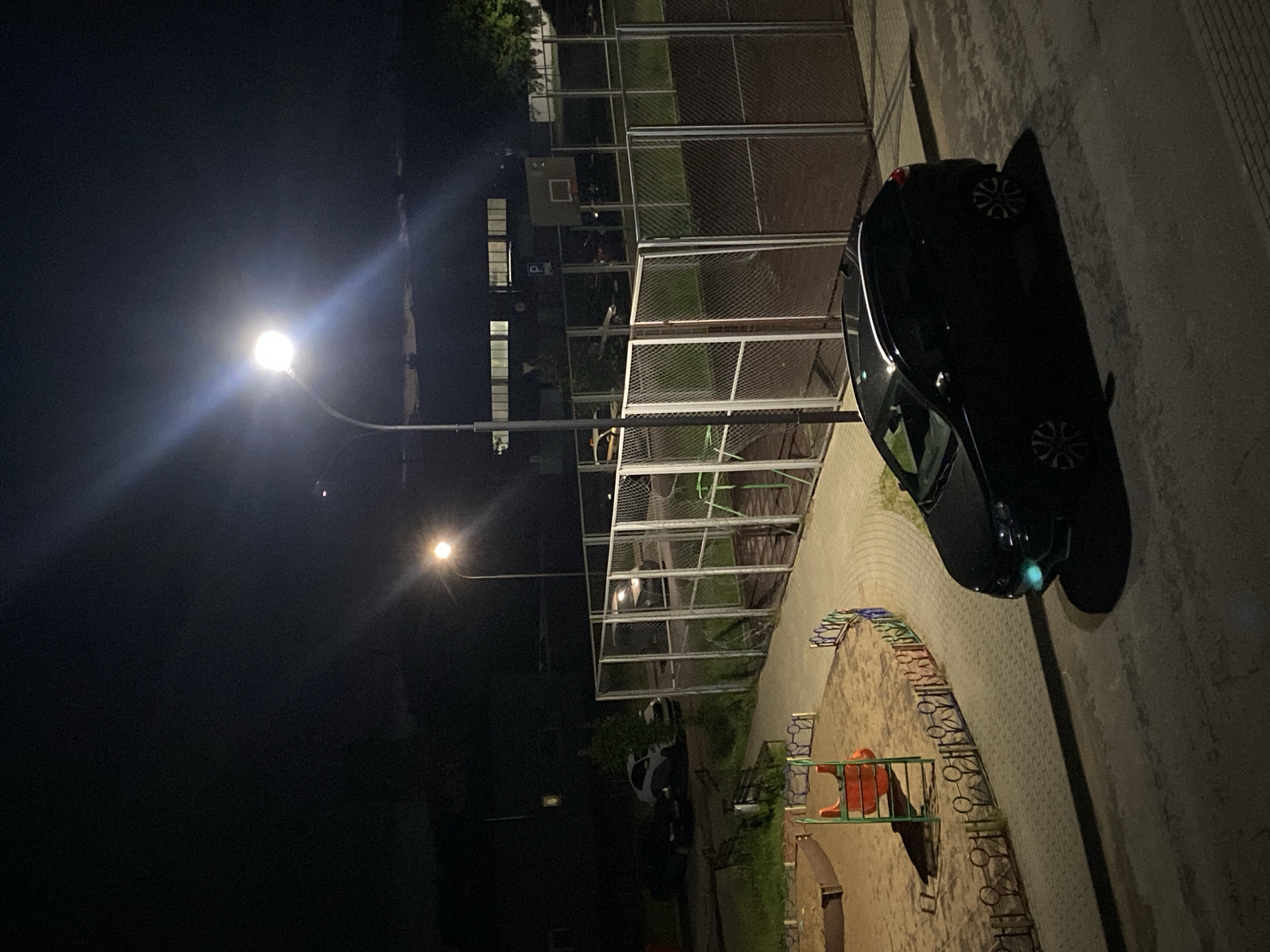 The image depicts an outdoor scene at night in an urban environment, possibly a residential complex, with artificial lighting from a streetlamp. In the foreground, a black sedan is parked on an asphalt surface, casting a long shadow. To the left, a children's playground with a sandbox and a red and green slide is visible, bordered by a decorative fence. Behind the playground, a paved walkway curves towards a fenced sports court, which appears to be a basketball court based on a backboard and hoop. Several other vehicles are parked in the background, near a building with illuminated windows. The overall atmosphere is one of quiet nighttime activity in a community space.