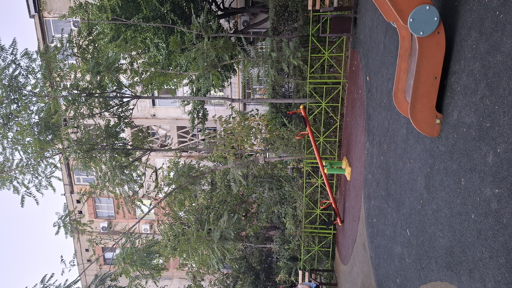 The image depicts a children's playground located in Shymkent, Kazakhstan. In the foreground, a black rubberized surface surrounds playground equipment. A bright orange seesaw with yellow and green accents is centrally positioned. To the right, the base of an orange slide is visible. A green metal fence encloses the playground area, separating it from a path and benches. Behind the fence, a dense arrangement of trees and bushes creates a natural backdrop. Beyond the foliage, a multi-story apartment building with light-colored facades and numerous windows dominates the background. The sky is overcast. A woman is seated on a bench to the left of the frame.