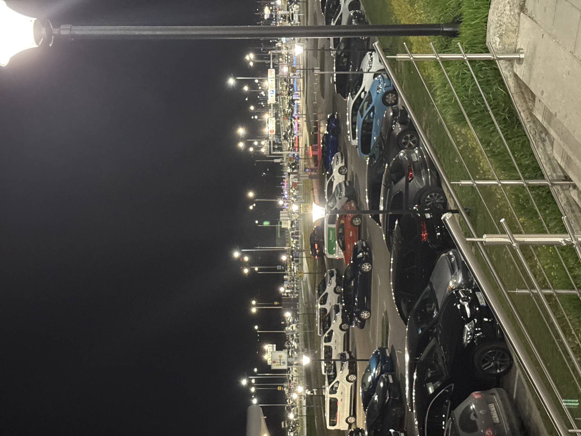 The image depicts a parking lot at night, densely packed with cars of various makes and models. The parking lot is illuminated by rows of lampposts extending into the distance. To the right of the parking lot, there's a retaining wall with a metal railing, suggesting an elevated viewpoint. Beyond the railing, there is vegetation. The scene gives the impression of a busy airport parking facility in Sofia, Bulgaria. The sky is dark, and the image captures the artificial lights reflecting off the vehicles.