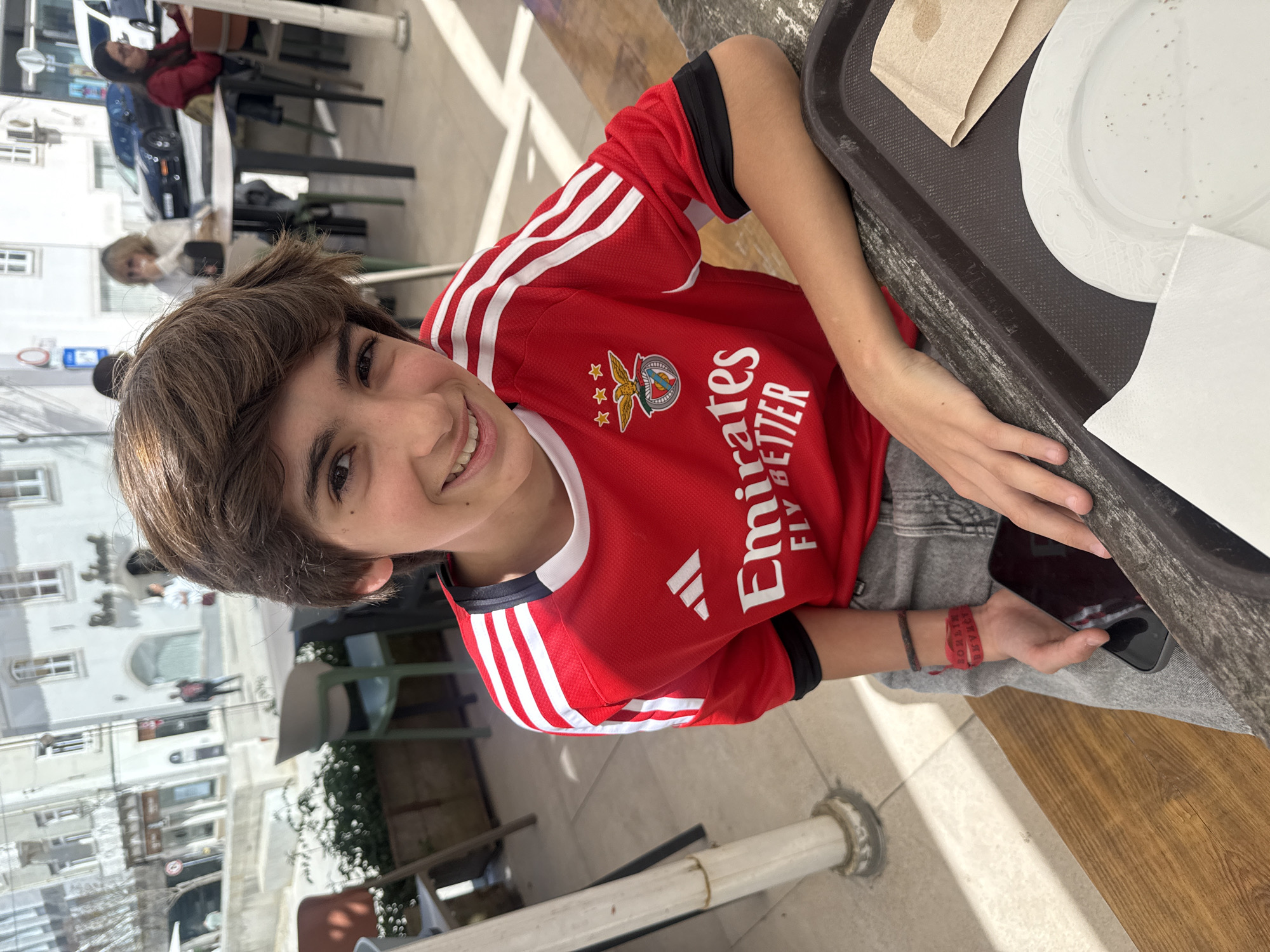 A medium shot, taken outdoors, shows a young person in a red soccer jersey with white stripes, seated at an outdoor table. The jersey has the emblem of a soccer club on it. The individual has medium-brown hair, light skin, and smiles slightly at the camera. They are holding a black smartphone in their right hand. A tray with a plate and napkins is visible at the right of the frame. The background shows a street scene with buildings and other people seated at outdoor tables, suggesting a cafe or restaurant setting. The scene is in Torres Vedras, Portugal.